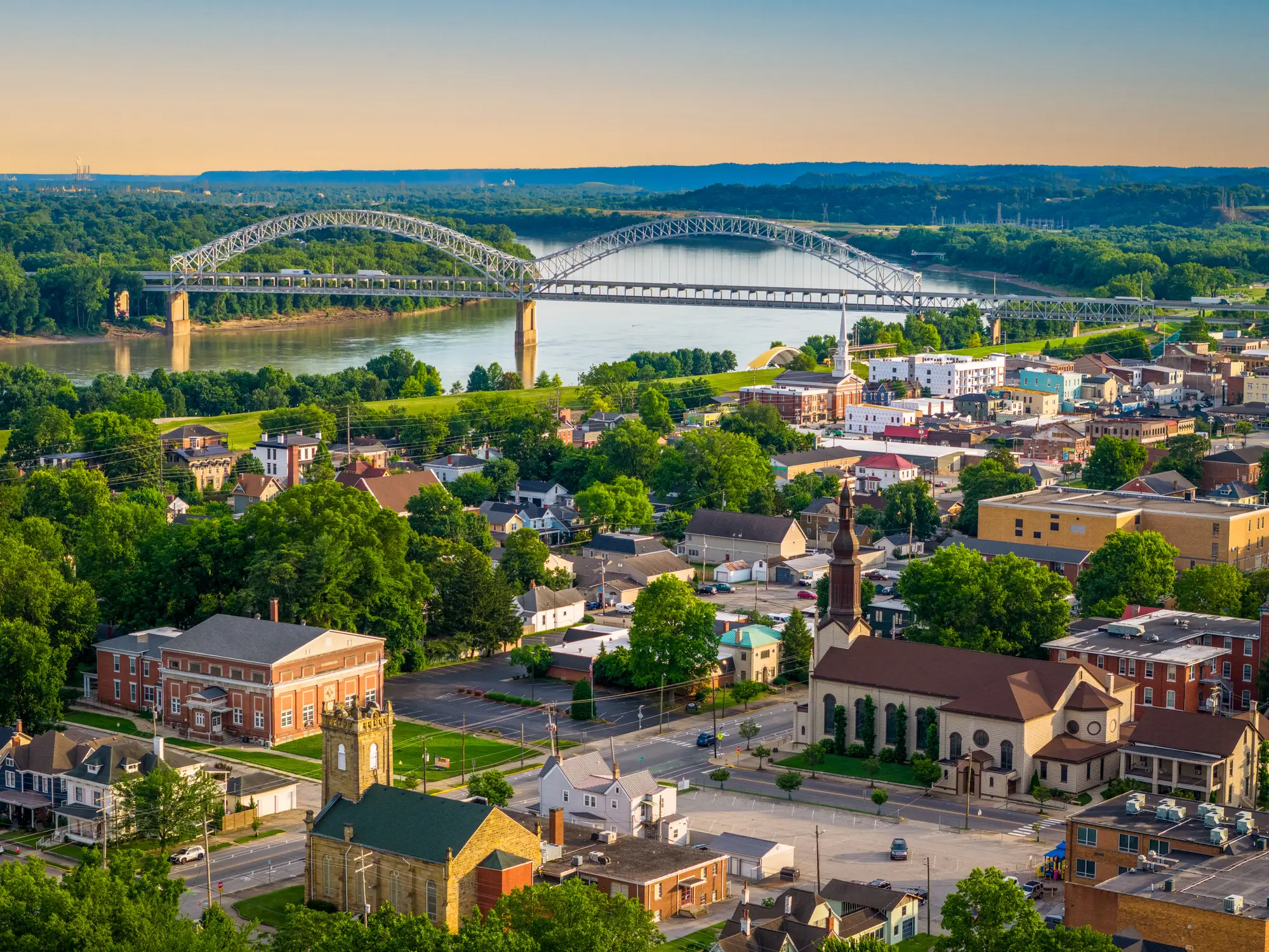 Aerial view of a riverfront town with steel arch bridges spanning a wide river at sunset.