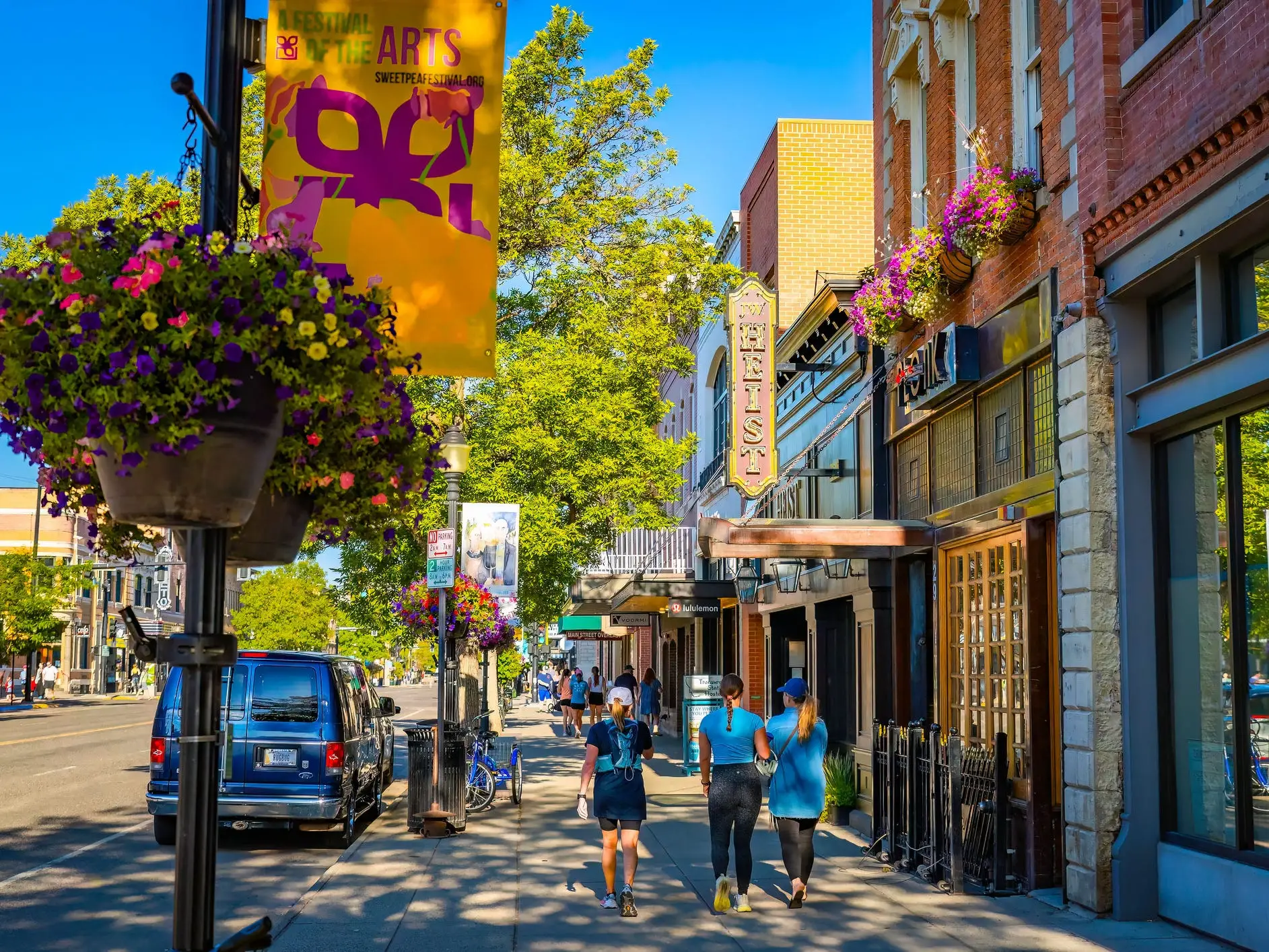 Pedestrians walk along a sunny downtown sidewalk past hanging flower baskets and an ELLEN theater sign.
