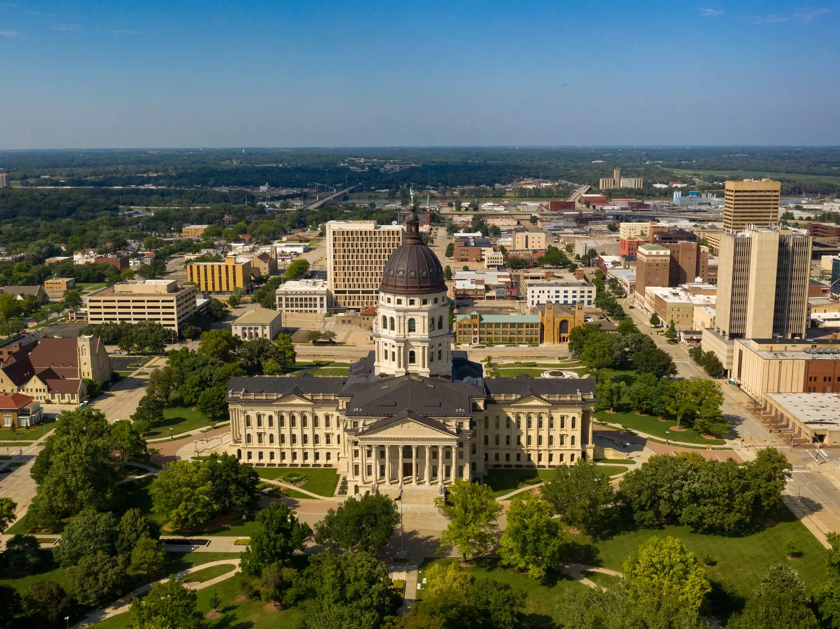 Aerial view of the Kansas State Capitol surrounded by downtown Topeka buildings and green tree-lined grounds.