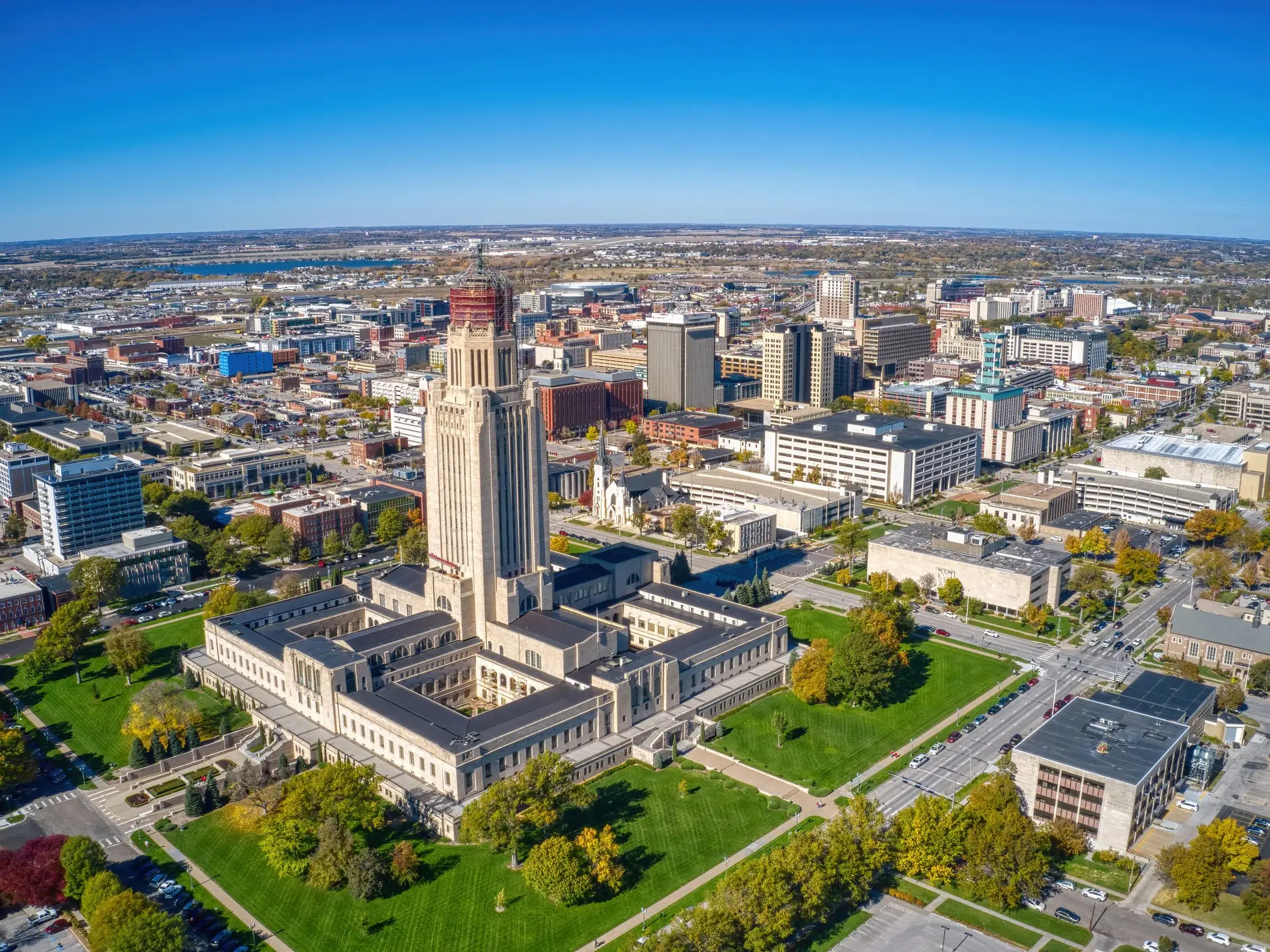 Aerial view of the Nebraska State Capitol surrounded by downtown Lincoln streets, buildings, lawns, and autumn trees.