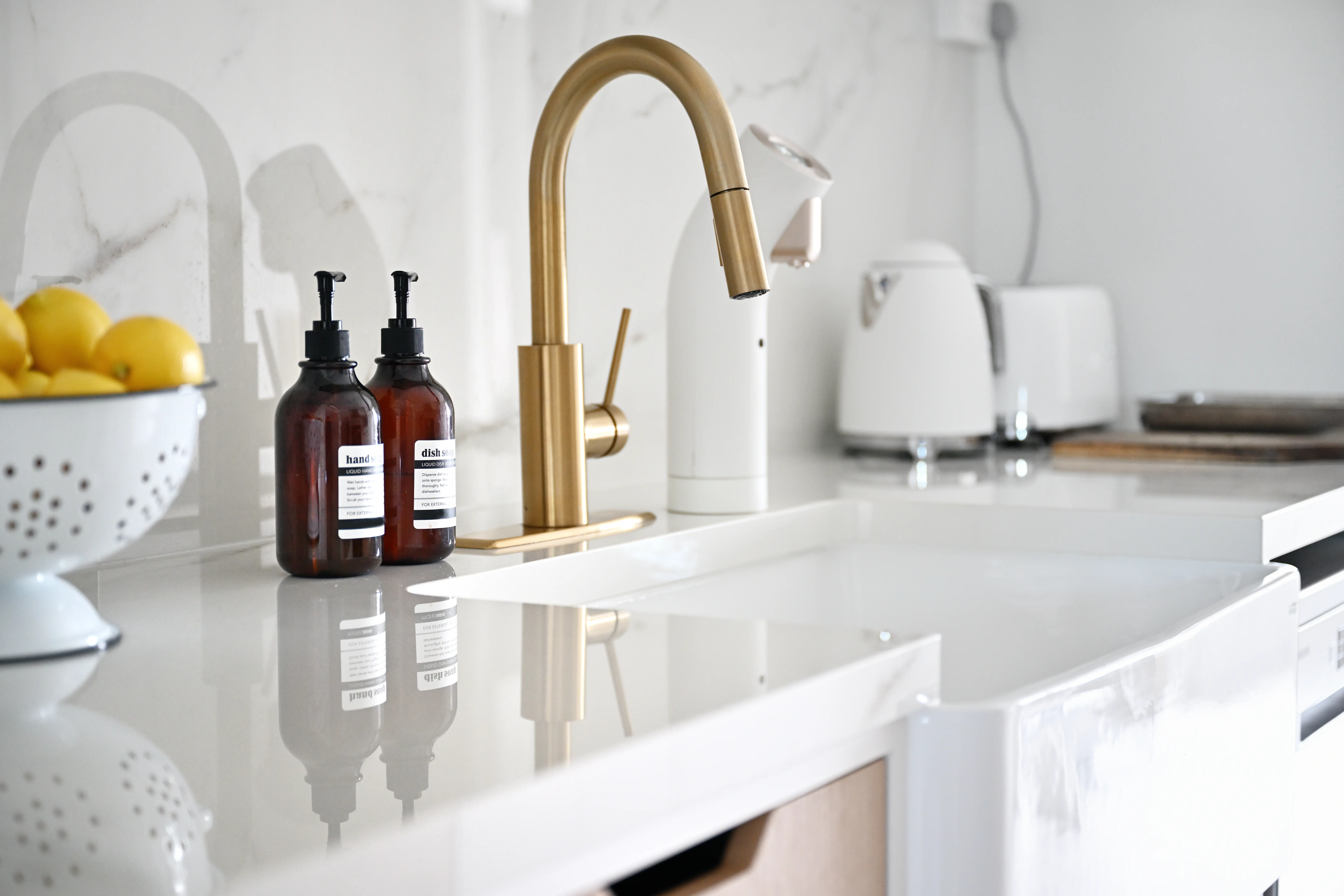 Bright kitchen counter with brass faucet, sink, soap dispensers, bowl of lemons, kettle, and toaster.