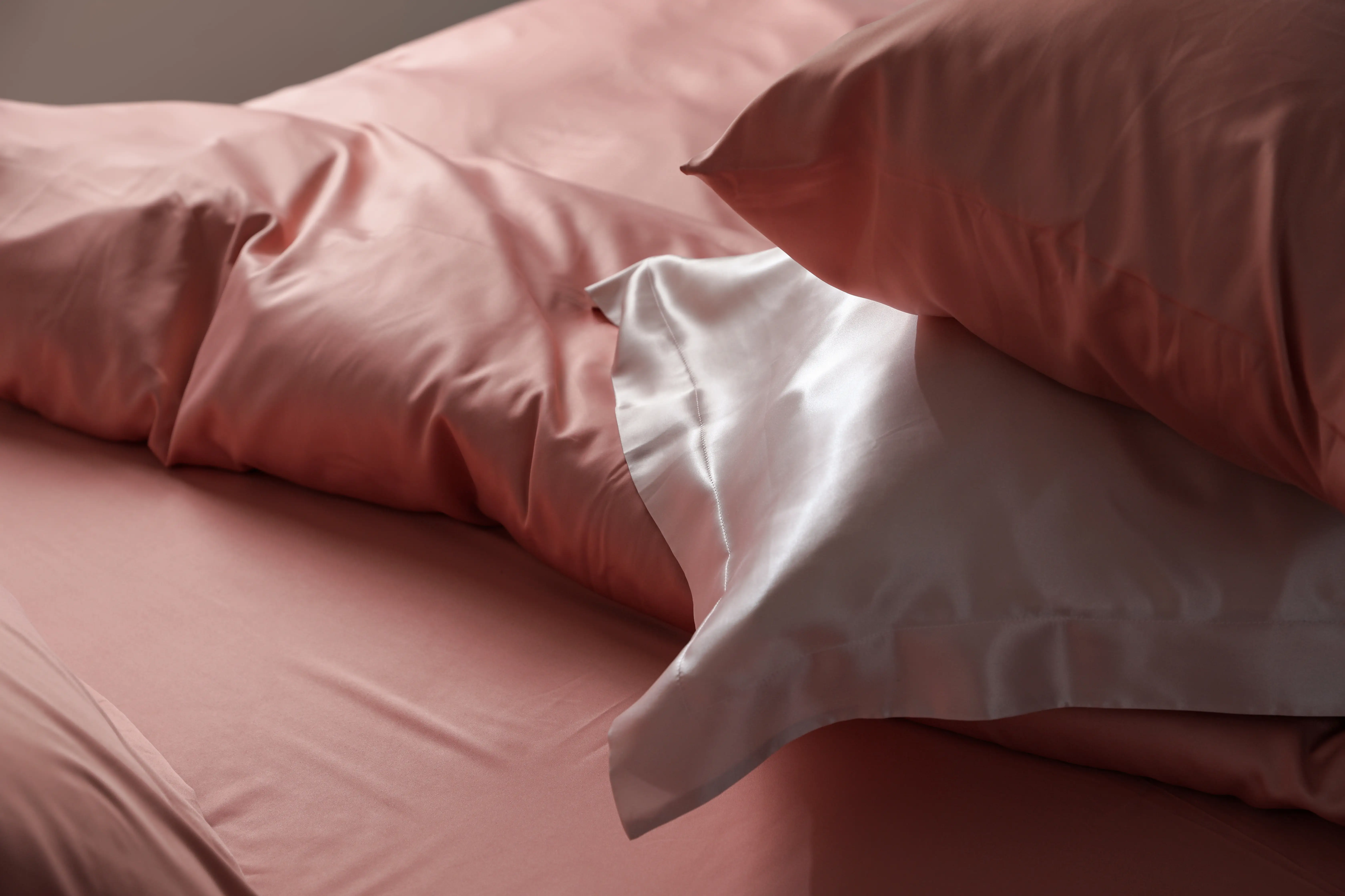 Pink bedding and pillows lie rumpled on a bed with a white pillowcase in soft natural light.