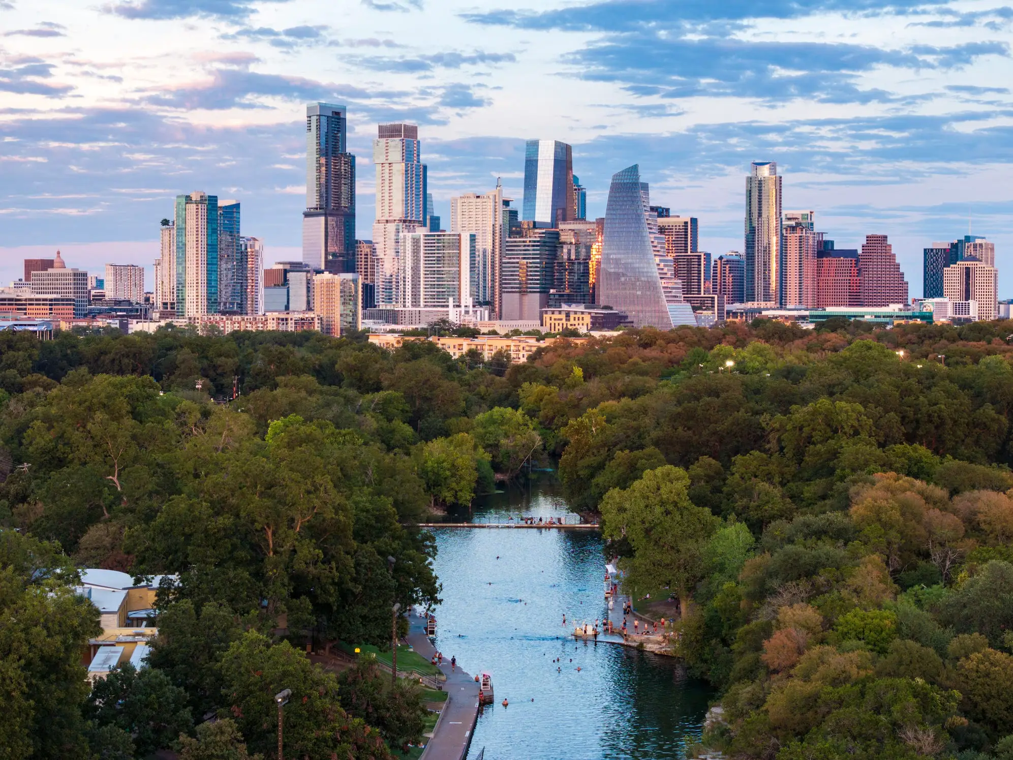 Downtown Austin skyline rises beyond trees and Barton Springs Pool at dusk.