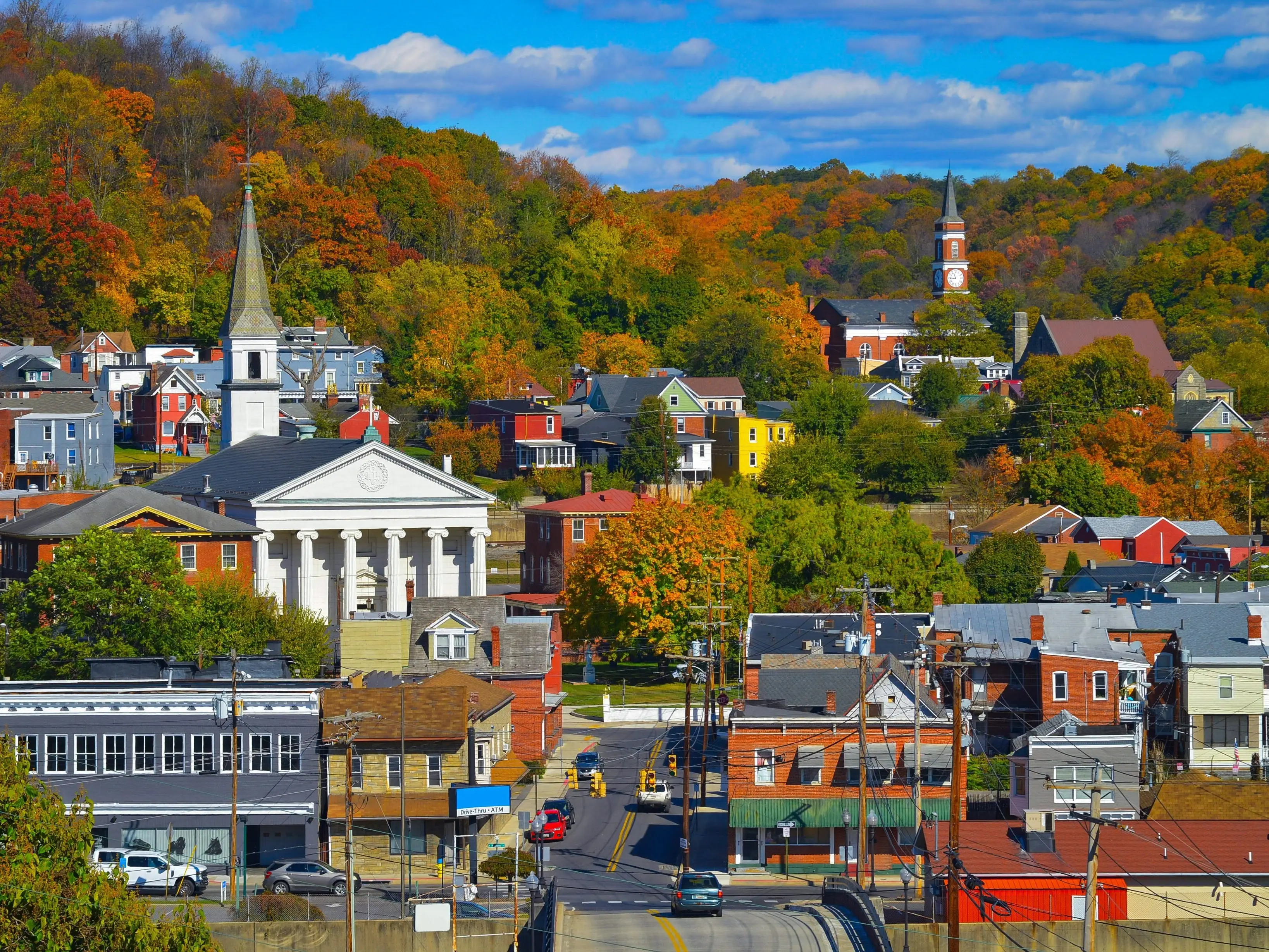 Small town streets and church steeples sit among colorful autumn hills under a blue sky.