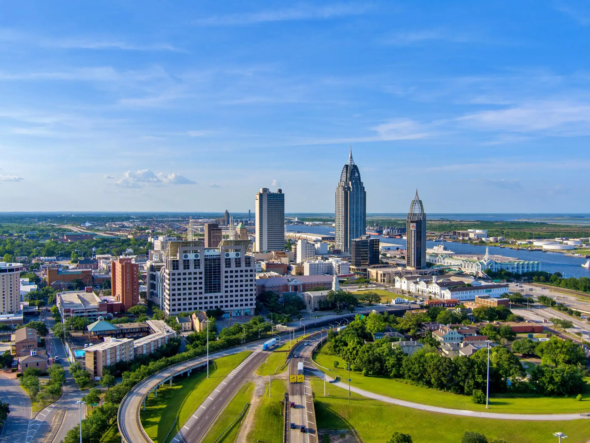 Downtown Mobile skyline rises beside the Mobile River with highways, greenery, and blue sky in the foreground.