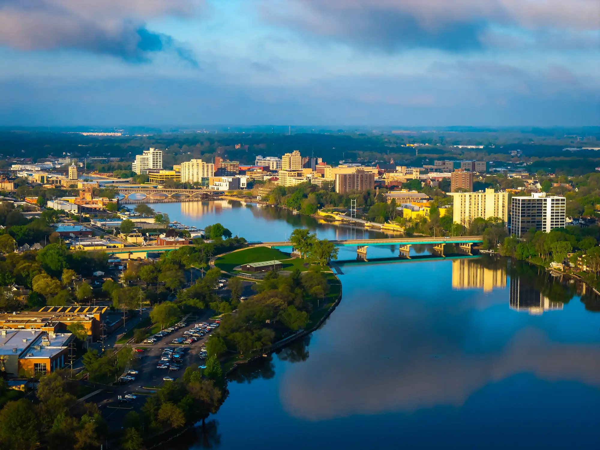 Aerial view of a downtown riverfront with bridges, high-rise buildings, trees, and cloud reflections on the water.