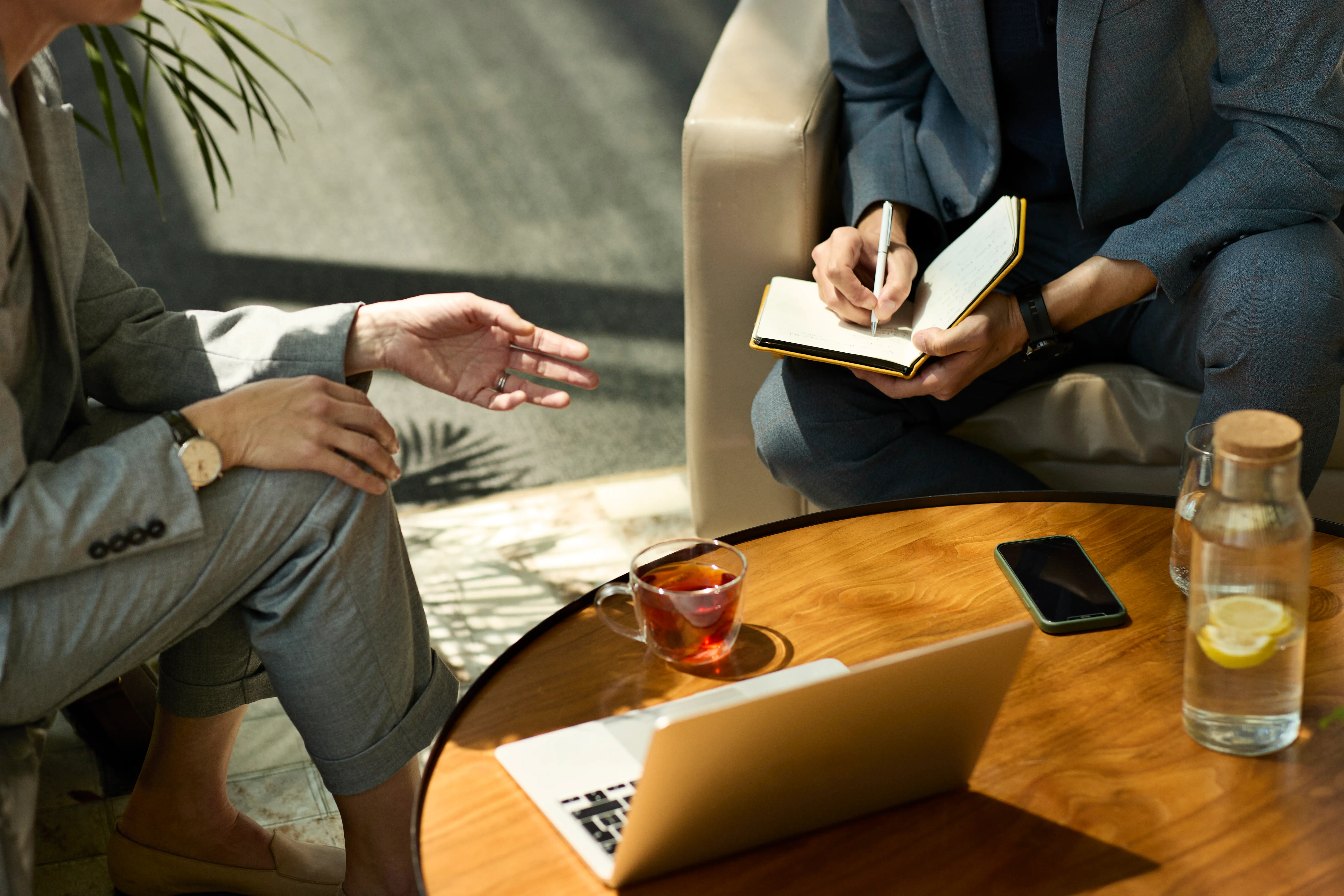 Two seated people discuss notes at a table with a laptop, phone, tea, and water.