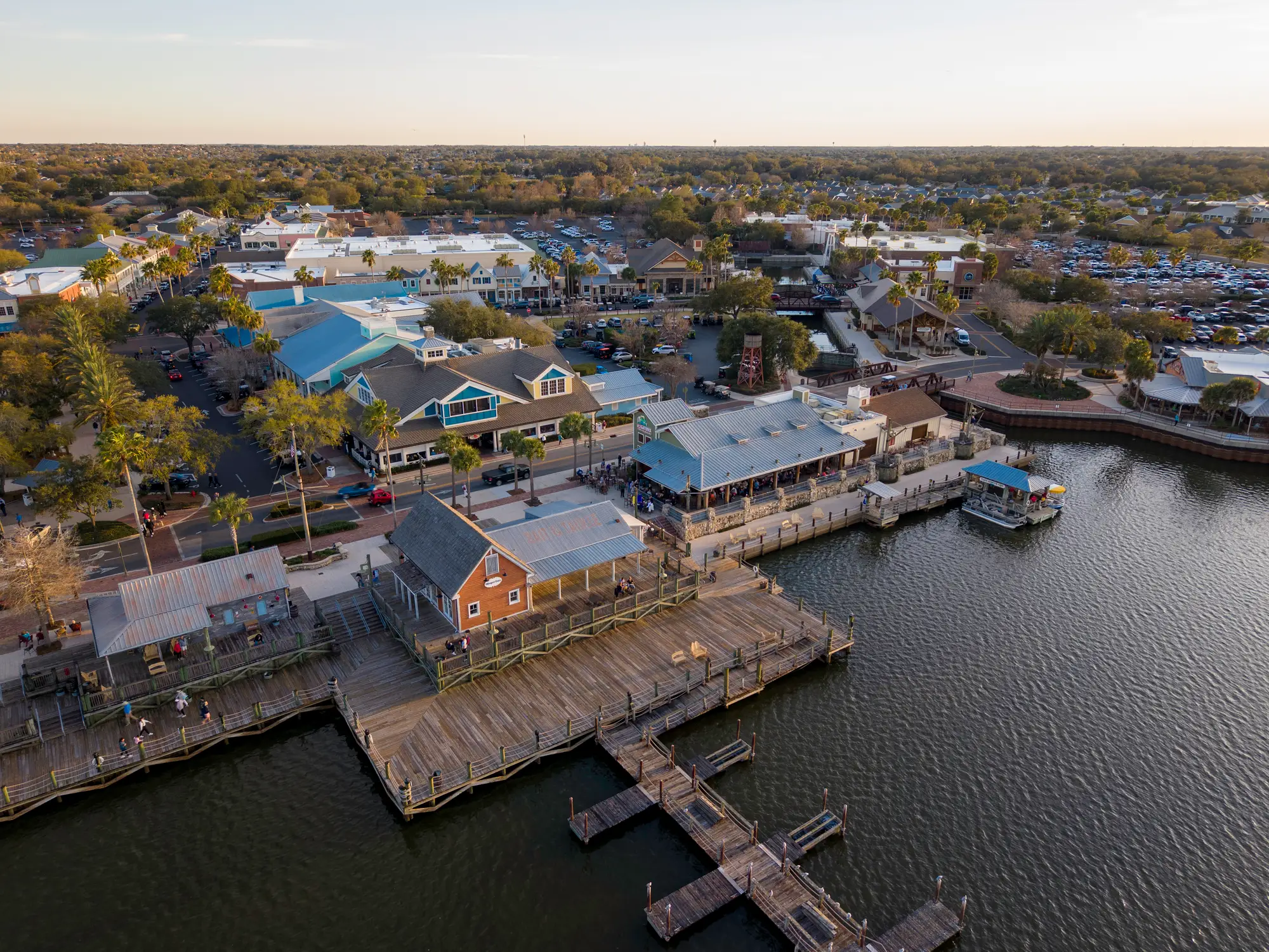 Aerial view of a waterfront shopping and dining district with boardwalks, docks, palm trees, and parked cars.