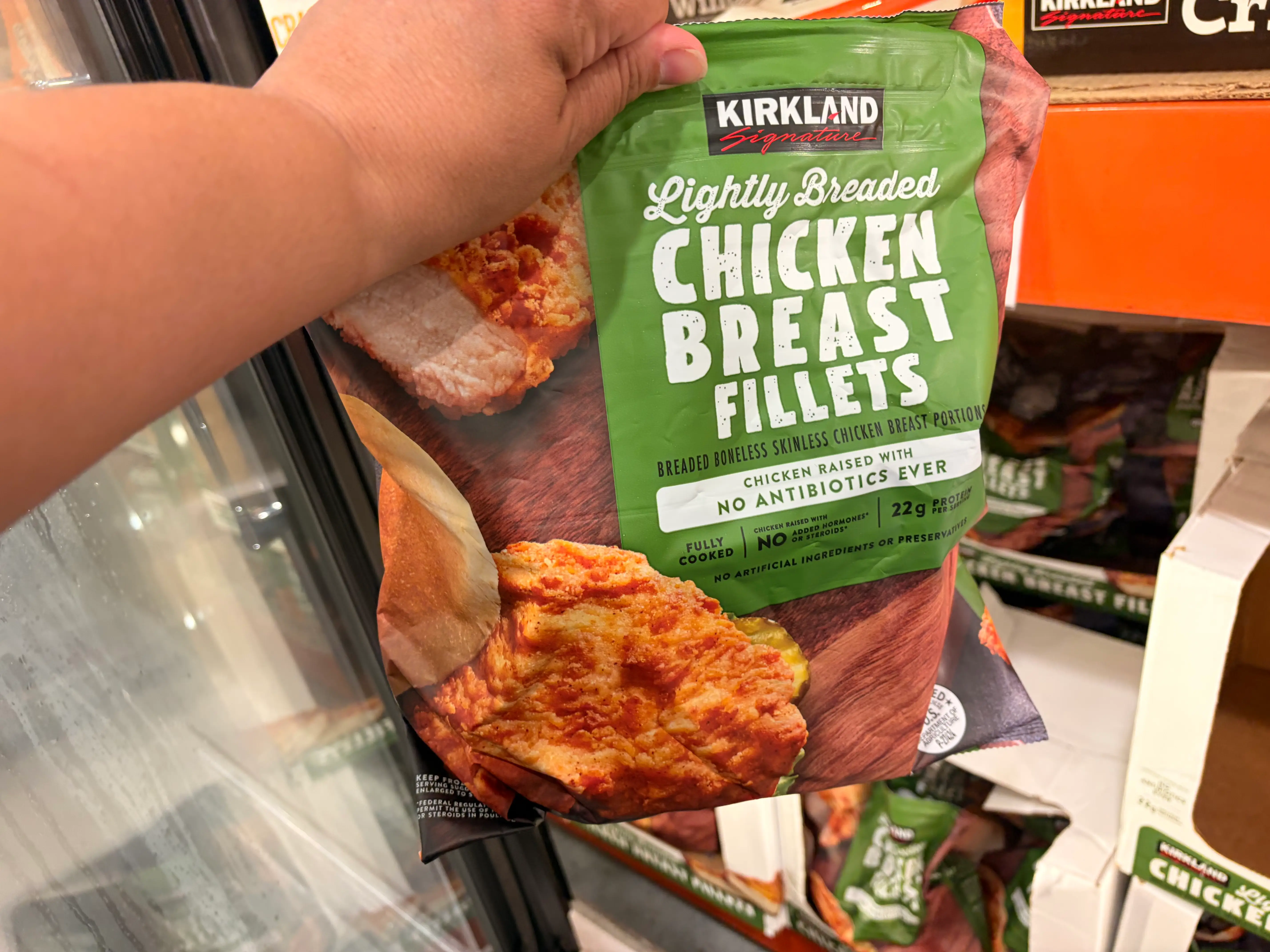 A hand holding a bag of Kirkland Signature lightly breaded chicken breast fillets.