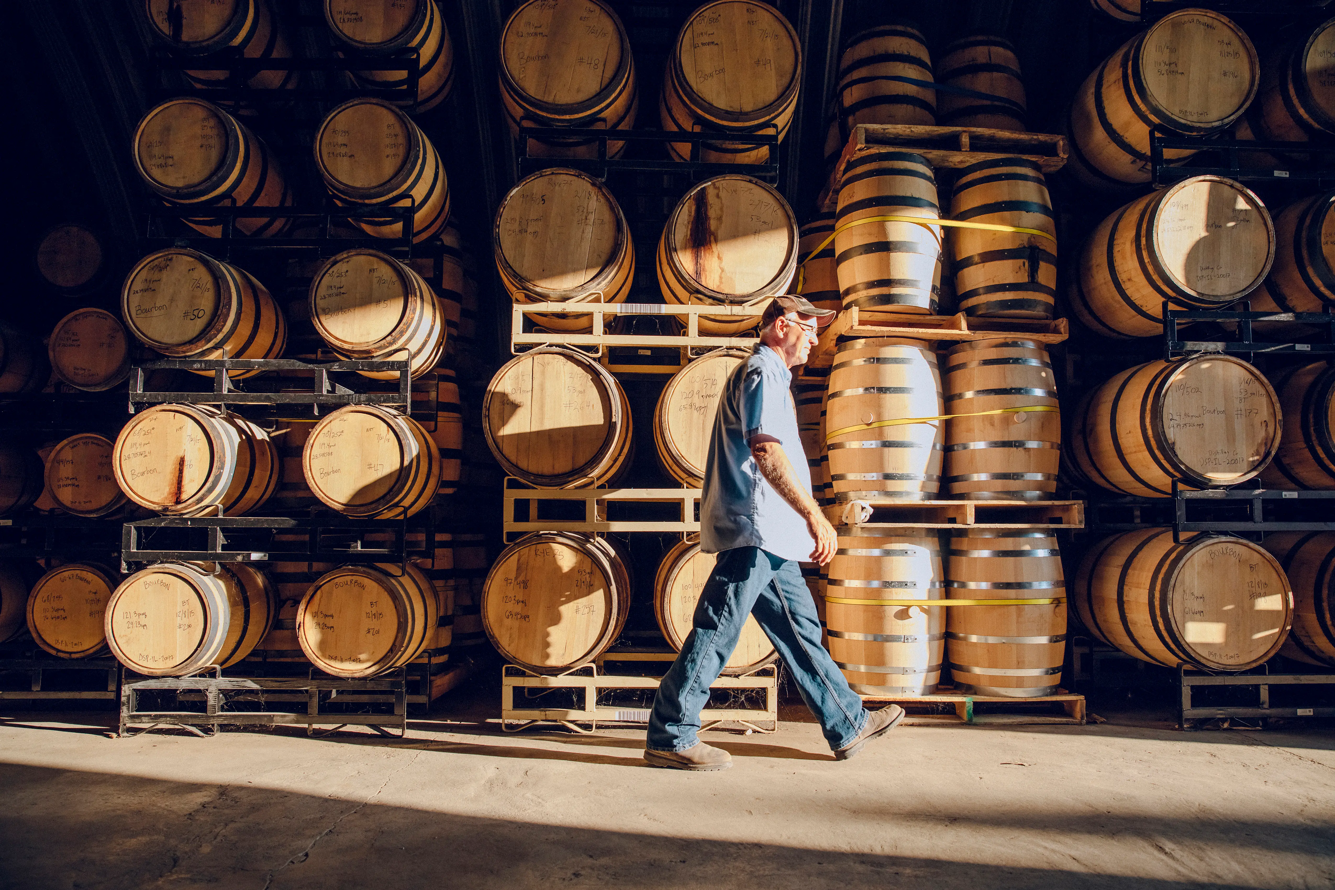 A person walks through a barrel storage area with rows of wooden casks stacked on metal racks.