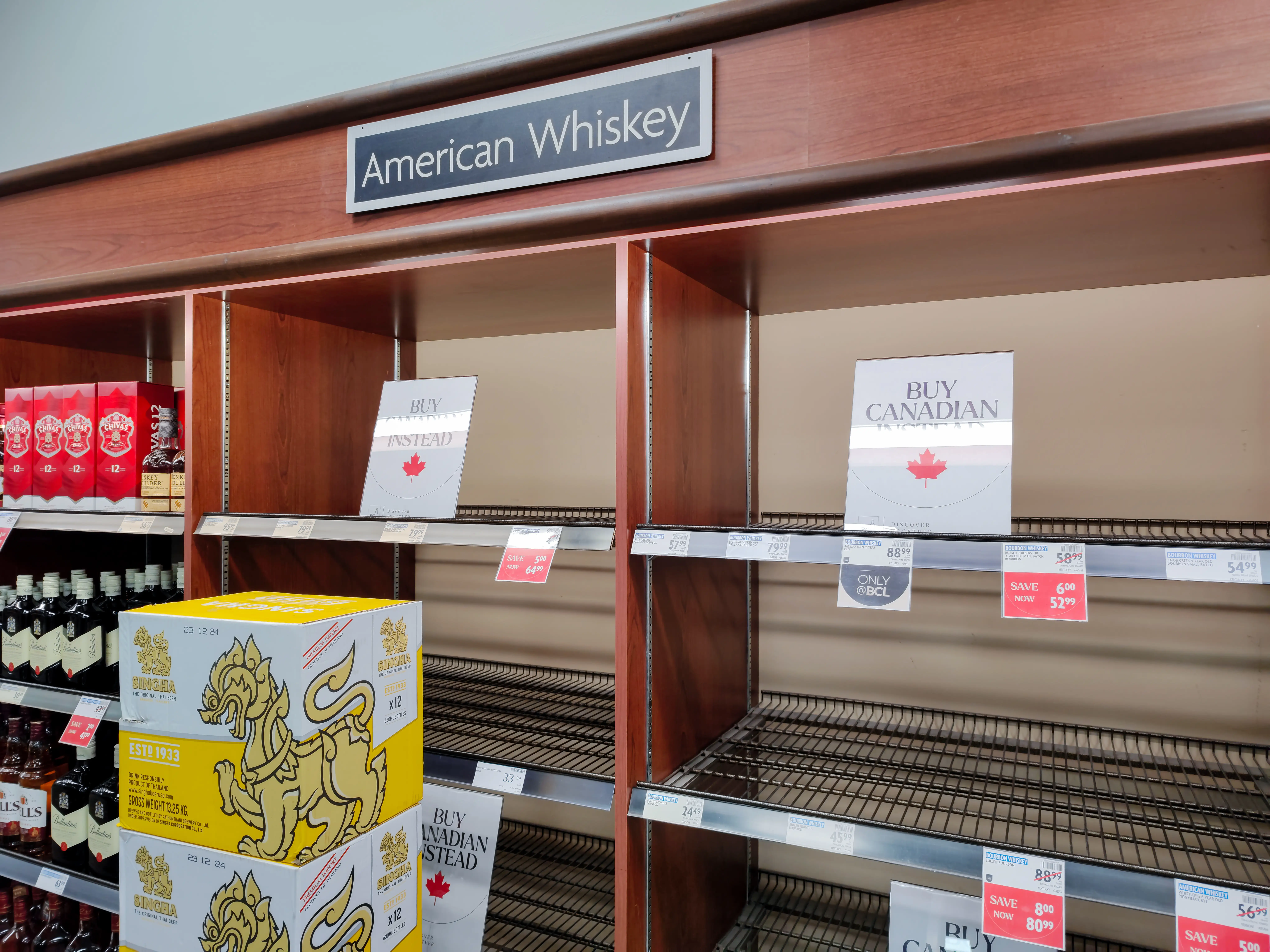 Mostly empty liquor store shelves under an “American Whiskey” sign display “BUY CANADIAN” notices and sale tags.