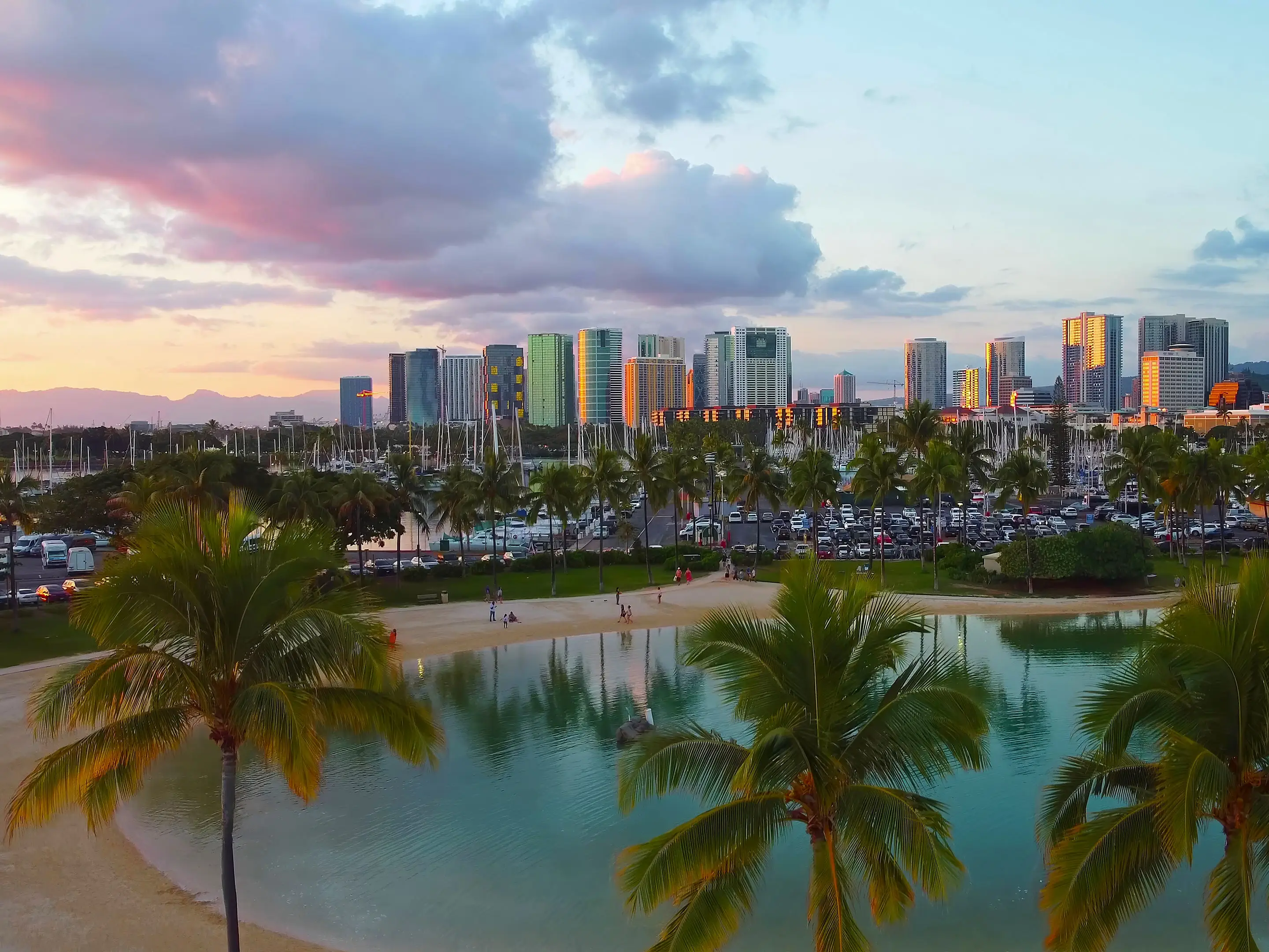 Palm trees frame a waterfront lagoon, marina, and high-rise skyline under colorful sunset clouds in Honolulu.