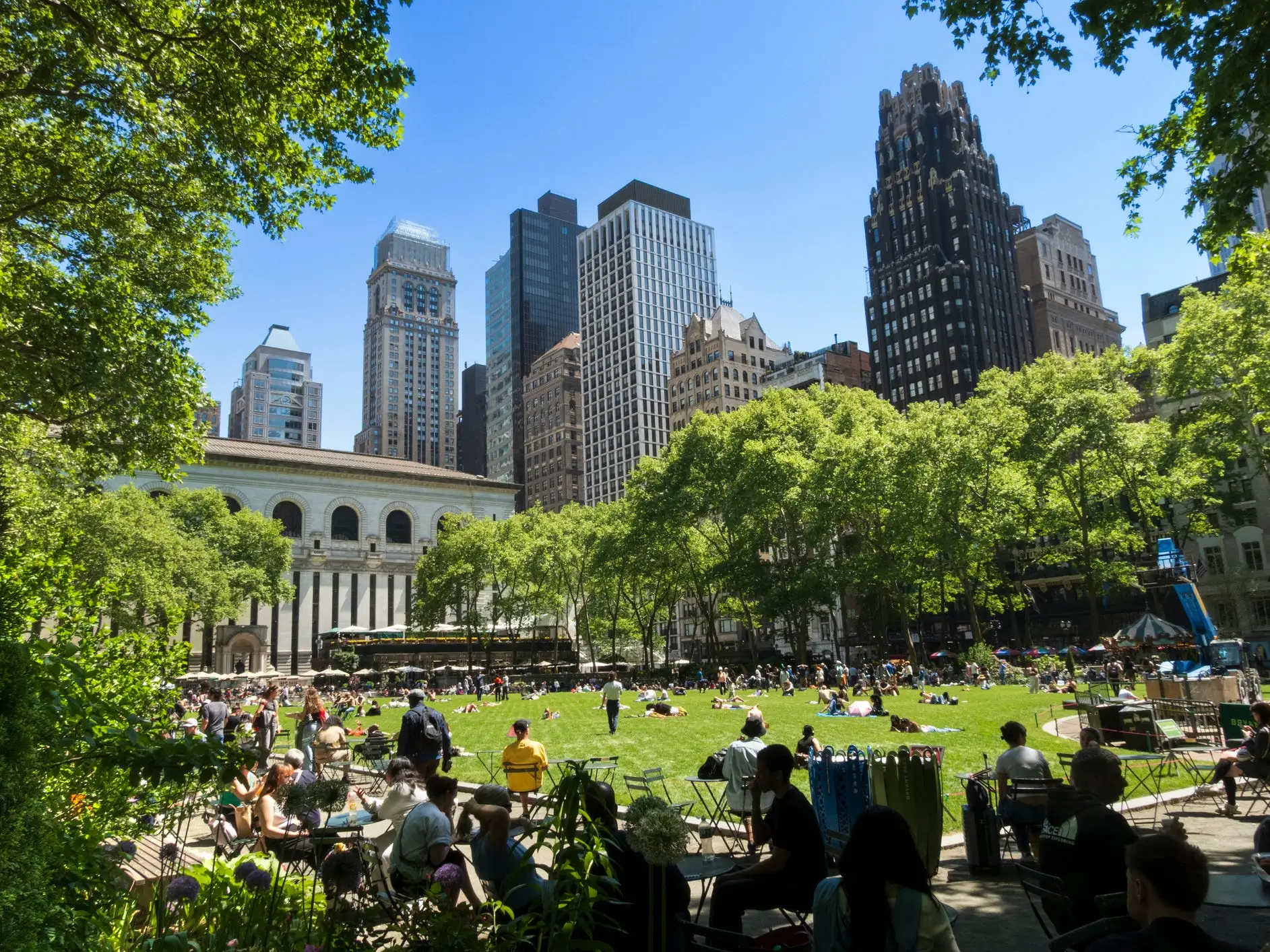 Crowds relax on Bryant Park lawn beside the New York Public Library and Midtown Manhattan skyscrapers on a sunny day.