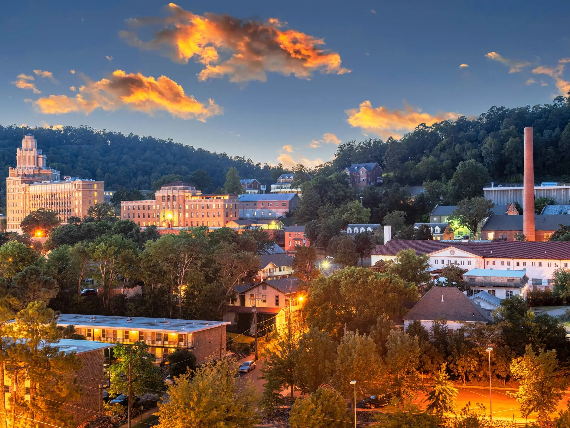 Sunset lights a forested hillside town with illuminated buildings, streets, and a tall brick smokestack.