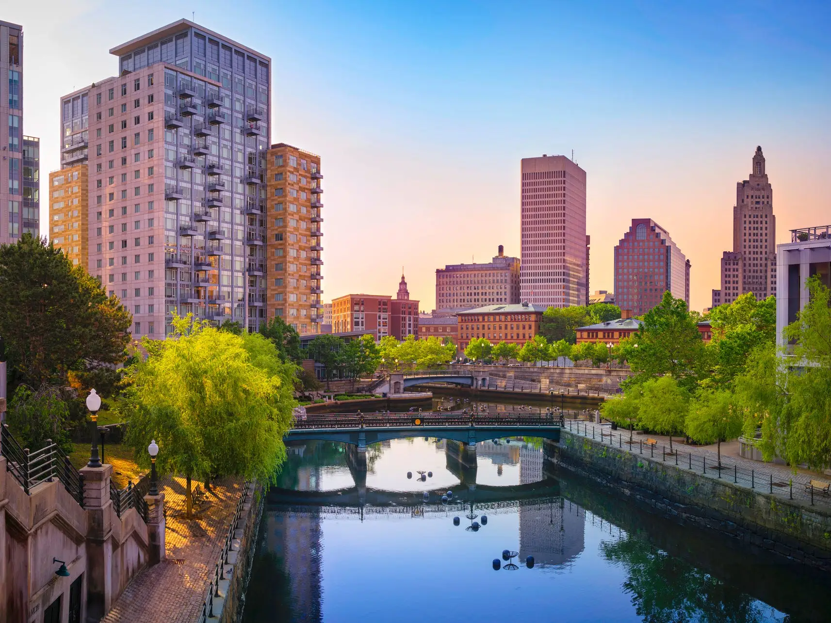 Downtown Providence skyline at sunset reflects in a calm river lined with trees, walkways, bridges, and modern buildings.