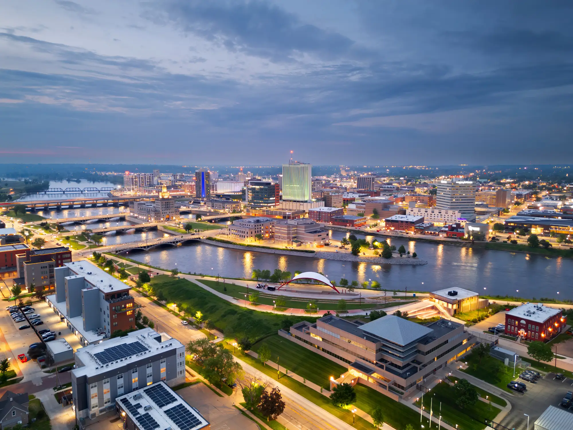 The downtown Cedar Rapids skyline glows at dusk along the Cedar River, with bridges, roads, and an amphitheater.