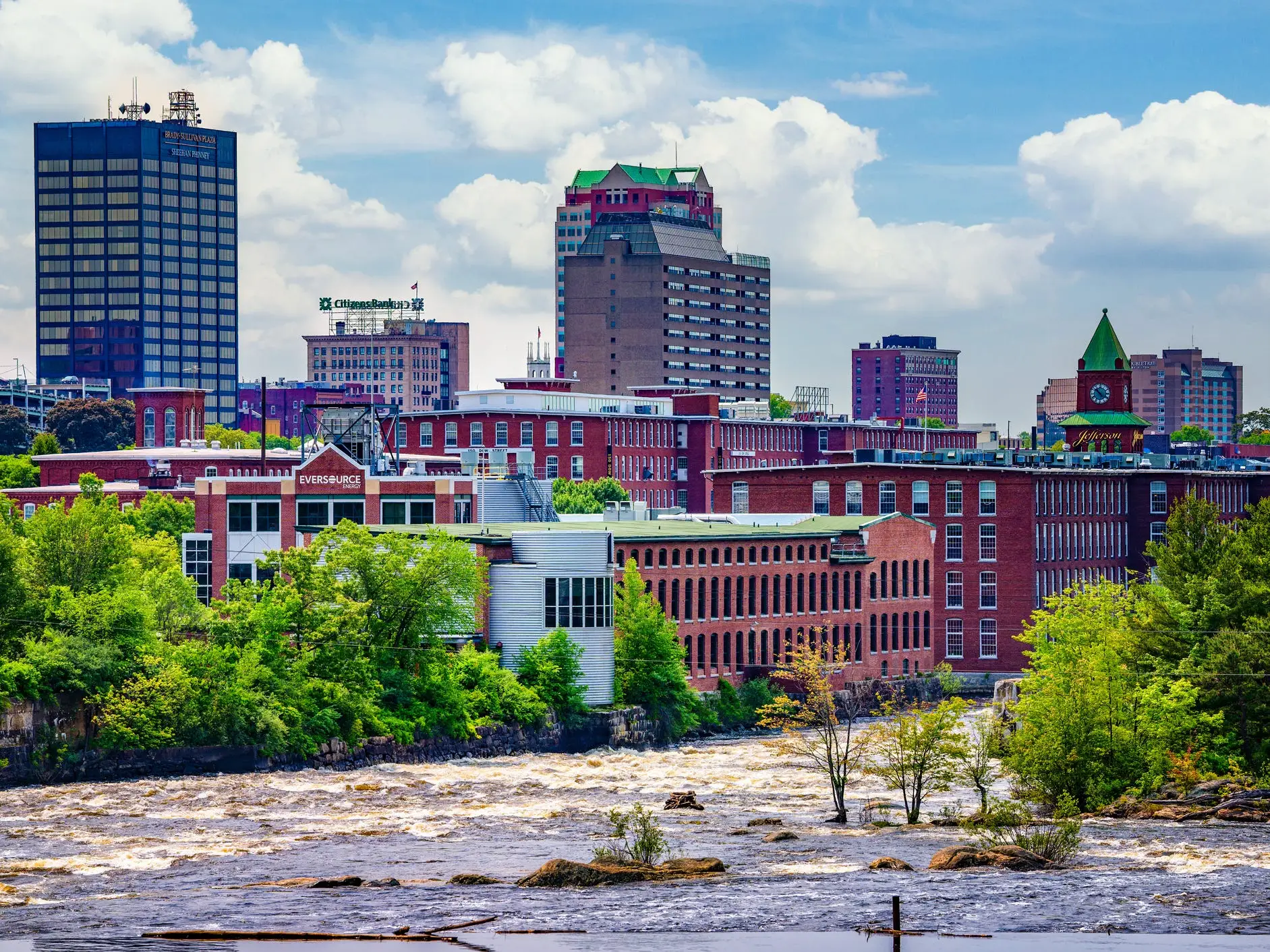 River rapids flow past brick mill buildings and downtown towers under a partly cloudy sky in Manchester, New Hampshire.
