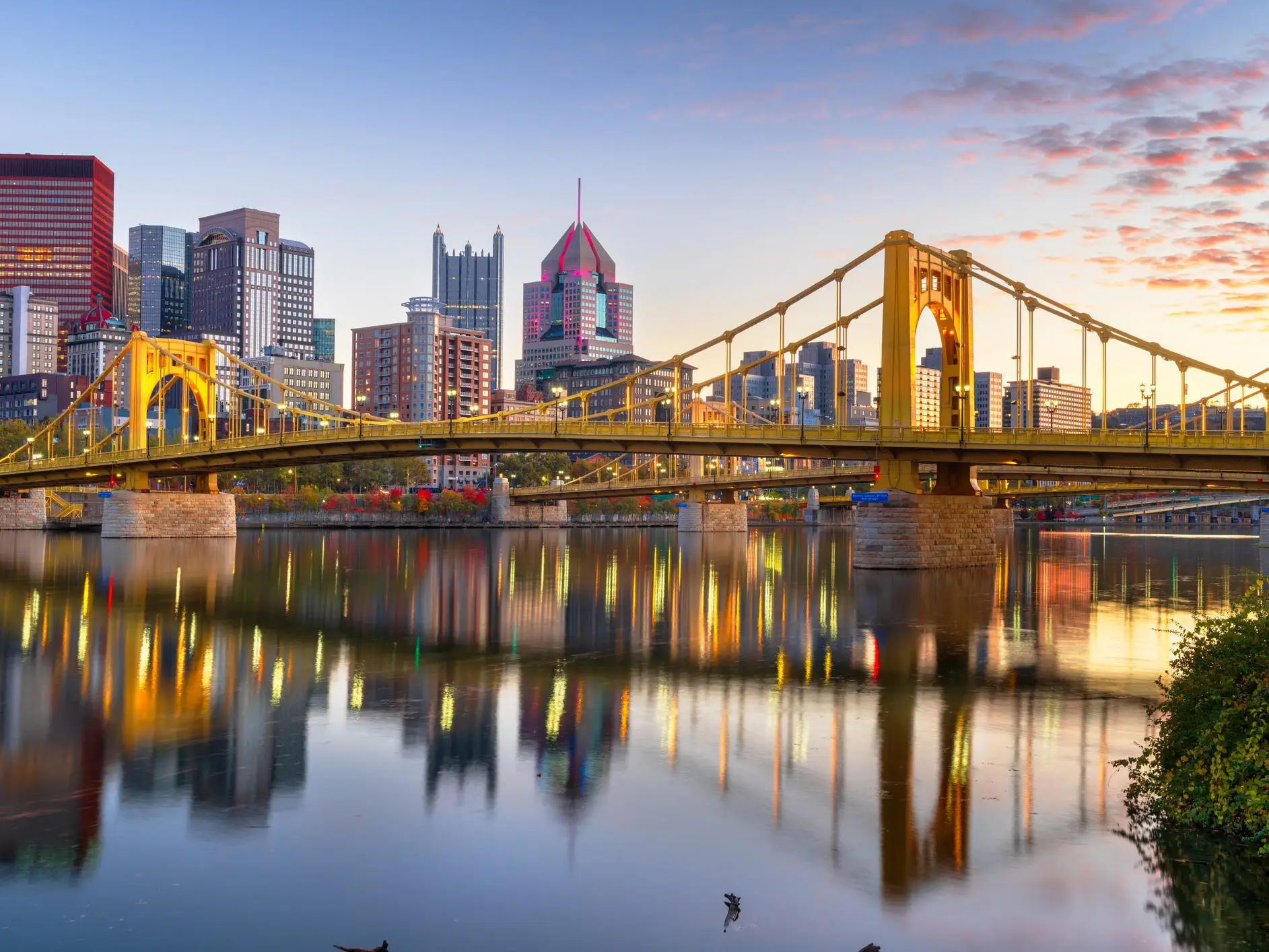 Pittsburgh skyline and a yellow suspension bridge reflect in a river at sunset.