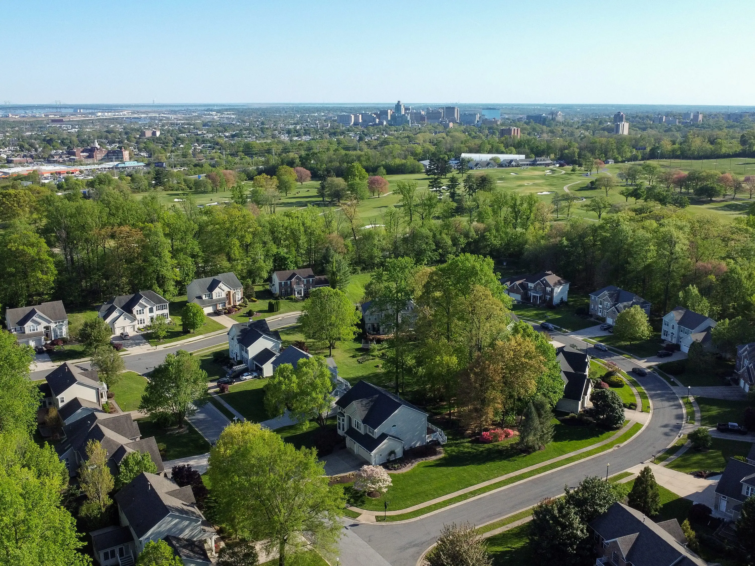 Aerial view of a tree-lined suburban neighborhood in Wilmington, Delaware, beside a golf course with a distant city skyline.