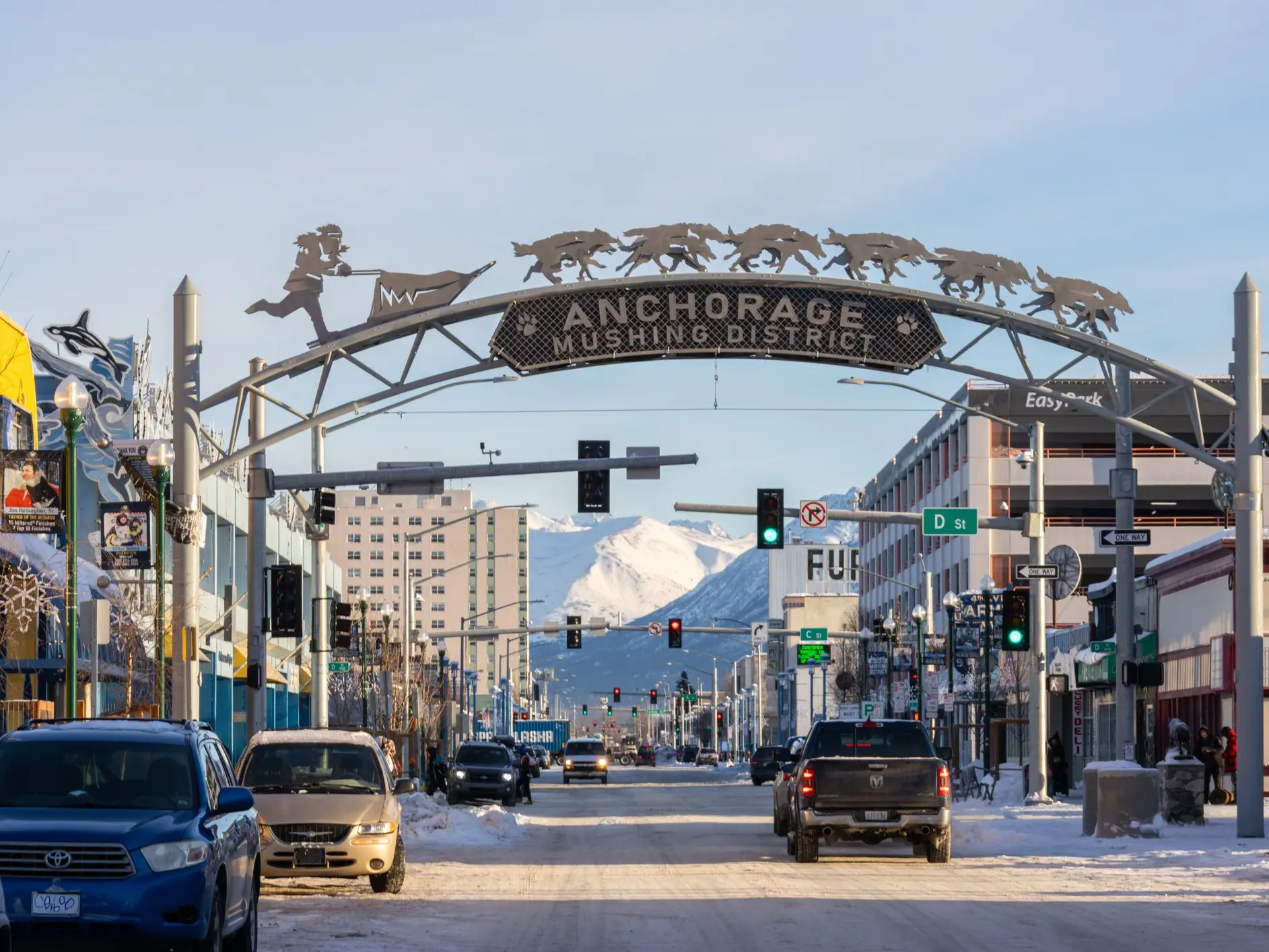 Snowy downtown Anchorage street beneath the Anchorage Mushing District arch, with cars, traffic lights, and mountains beyond.