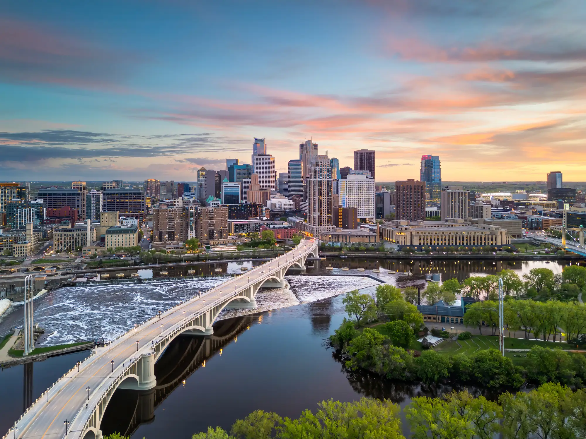 Downtown Minneapolis skyline rises behind the Mississippi River and an arched bridge at colorful sunset.