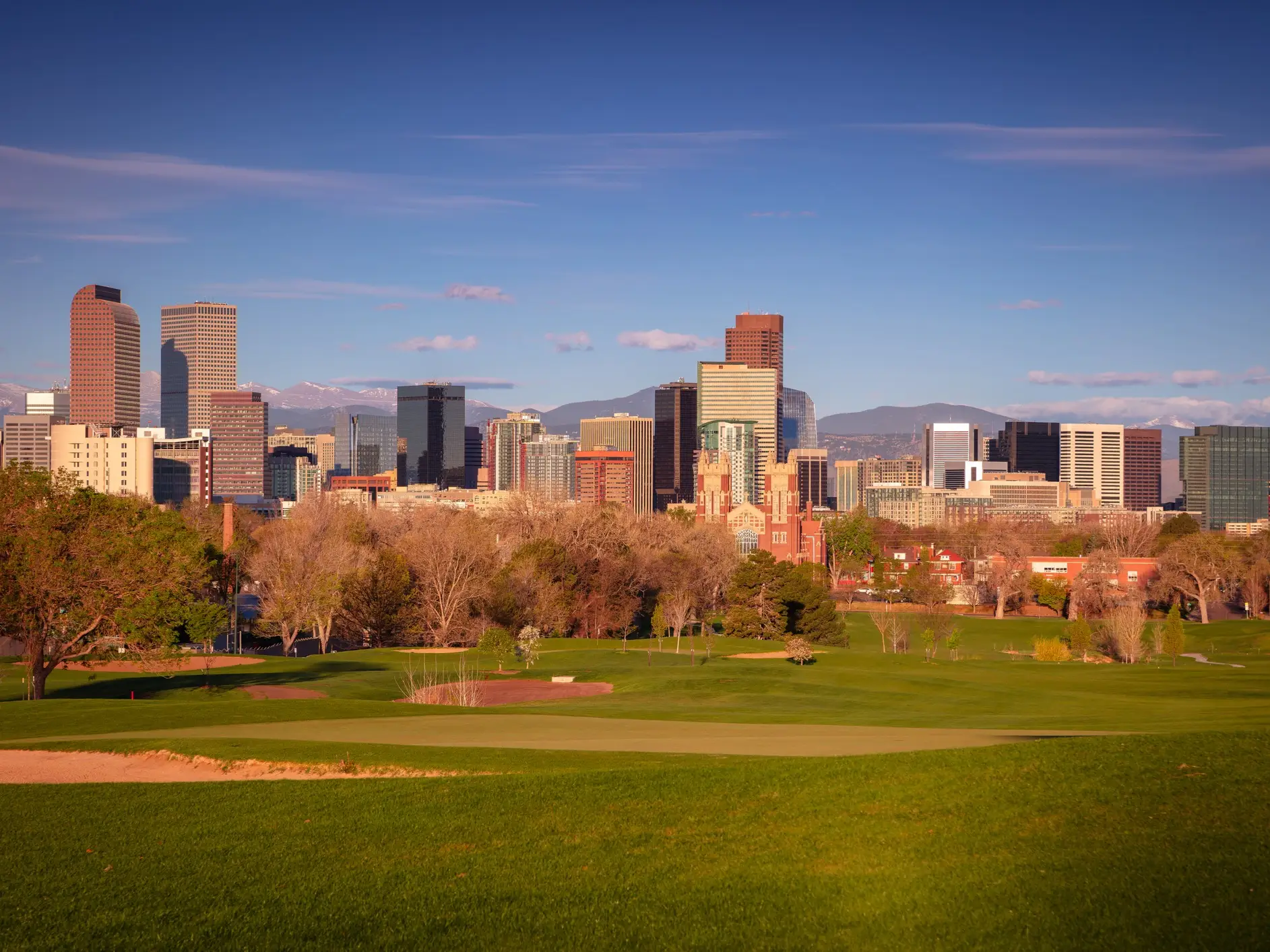 Denver skyline rises beyond a green golf course with trees and distant mountains under a clear blue sky.