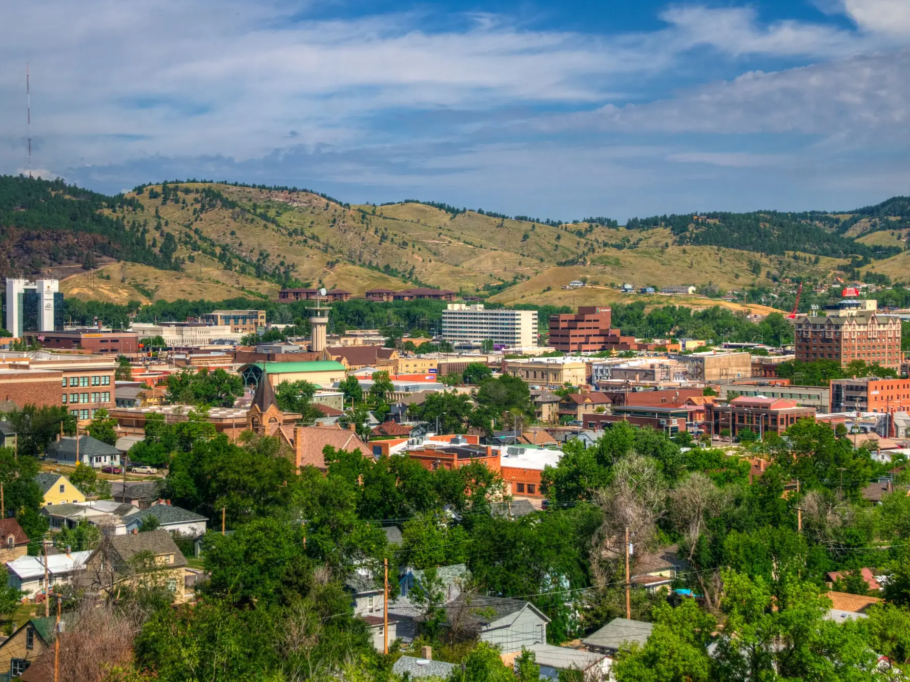 Downtown Rapid City buildings and homes sit beneath green Black Hills under a partly cloudy sky.