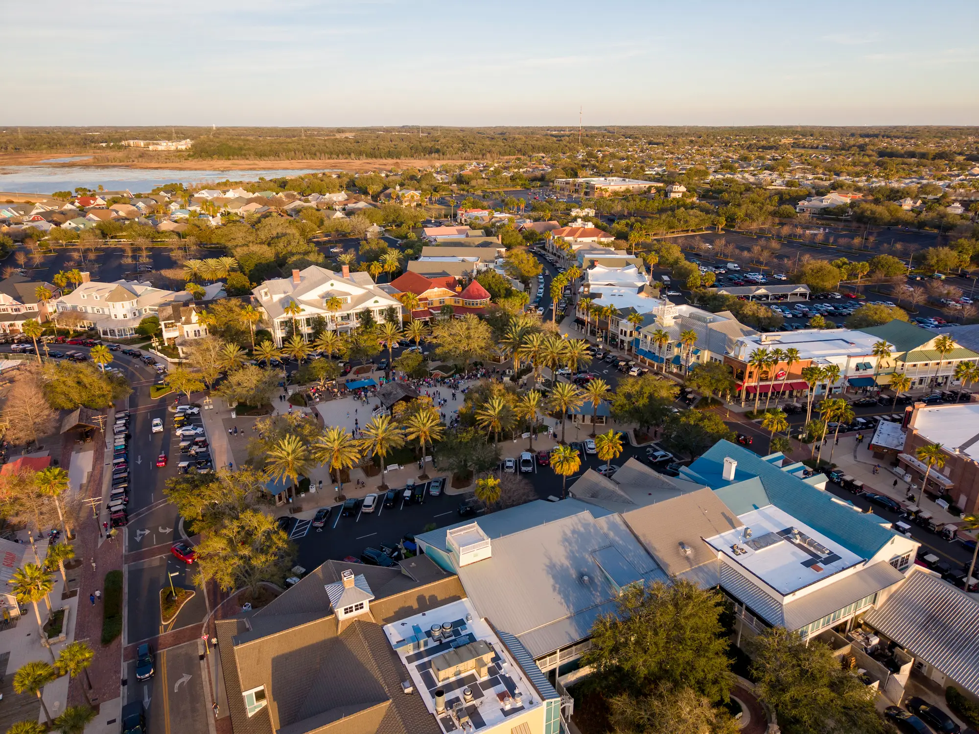 Aerial view of a palm-lined town center with shops, parking areas, homes, and a lake at sunset.