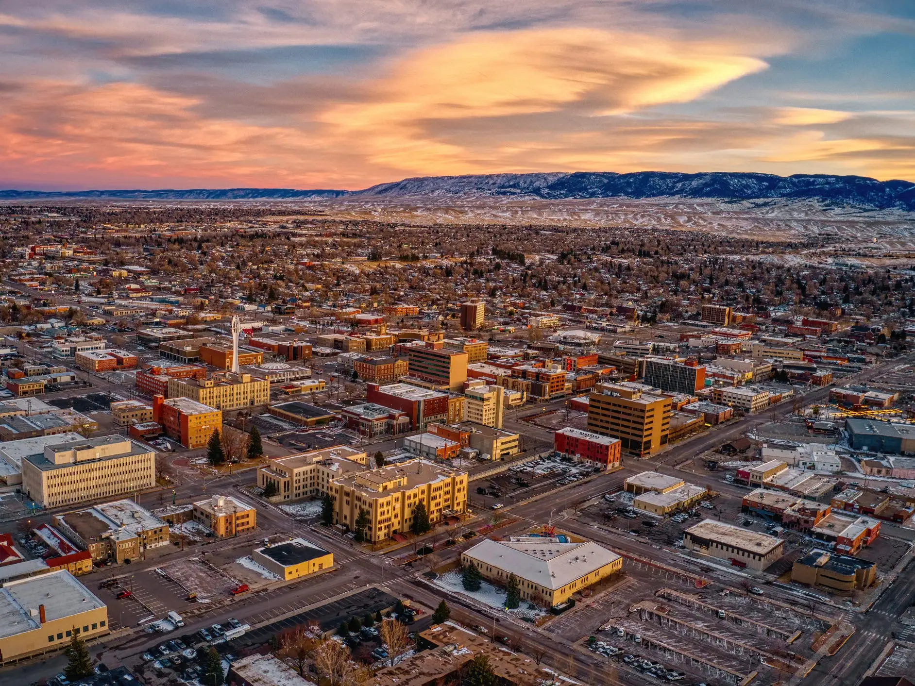 Aerial view of a snowy downtown area in Casper, Wyoming, with mountains in the distance under a colorful sunset sky.