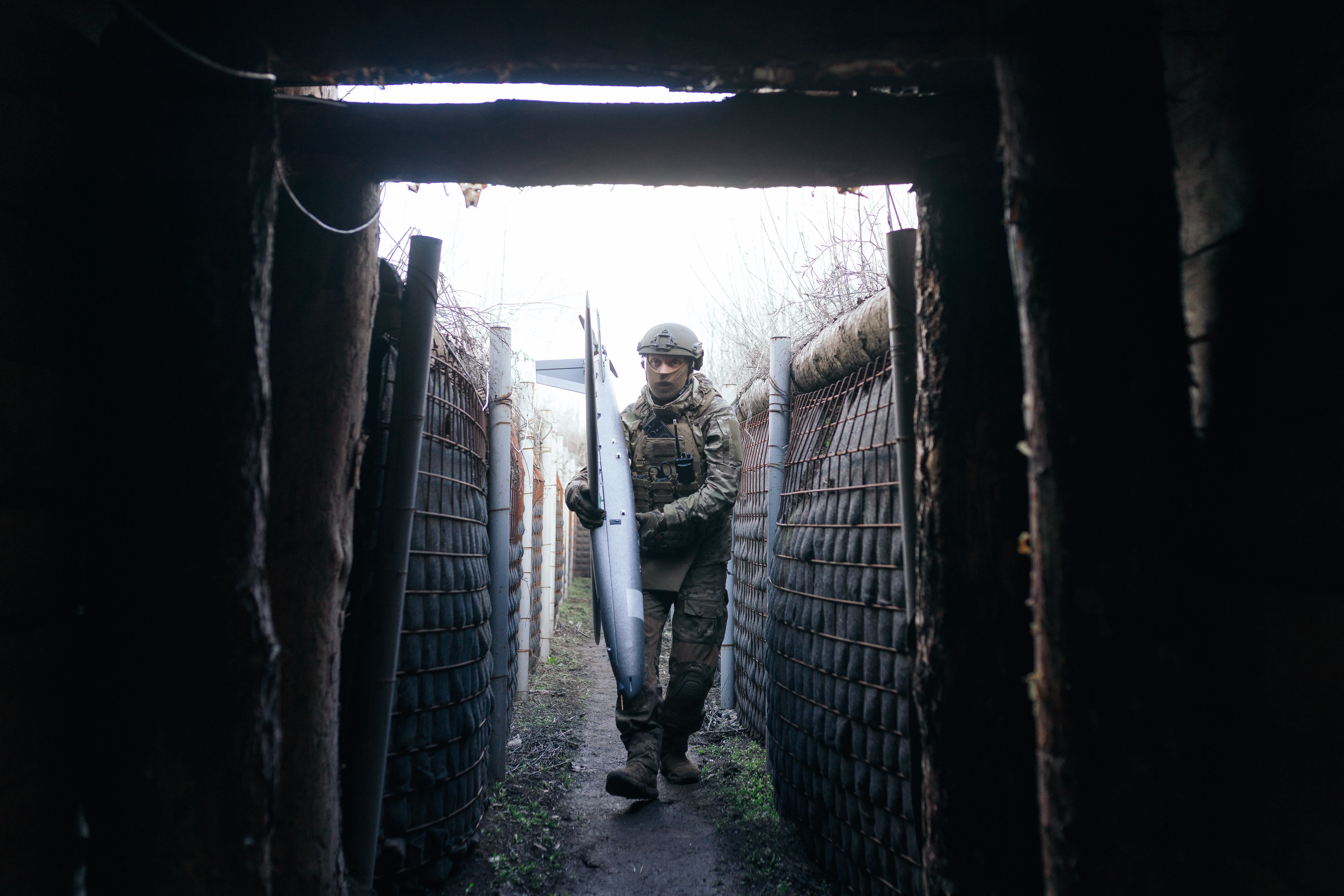 A uniformed soldier carrying a long object walks through a narrow reinforced trench toward a dark bunker entrance.