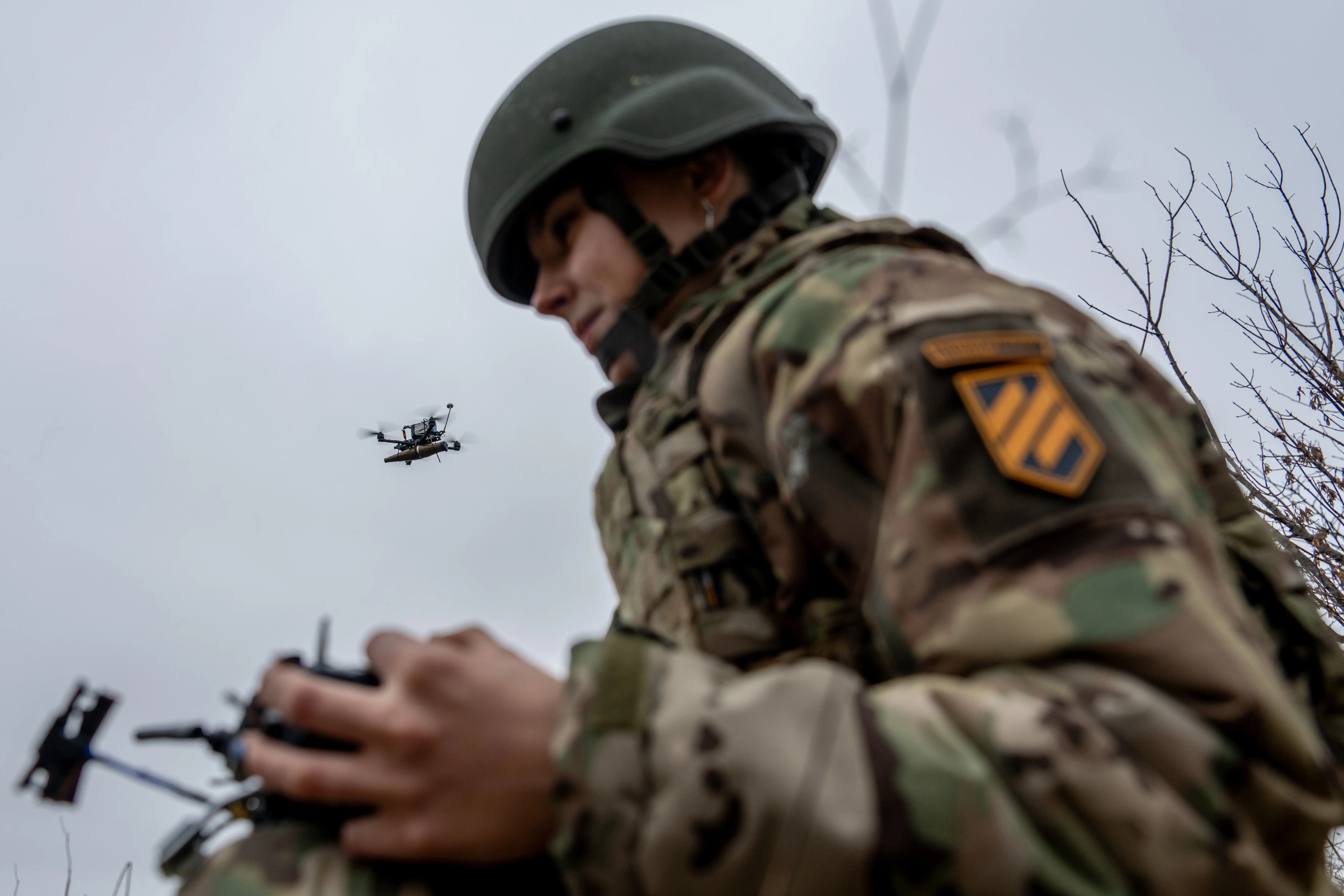 Camouflaged soldier in a helmet operates a drone controller while a small quadcopter flies nearby.