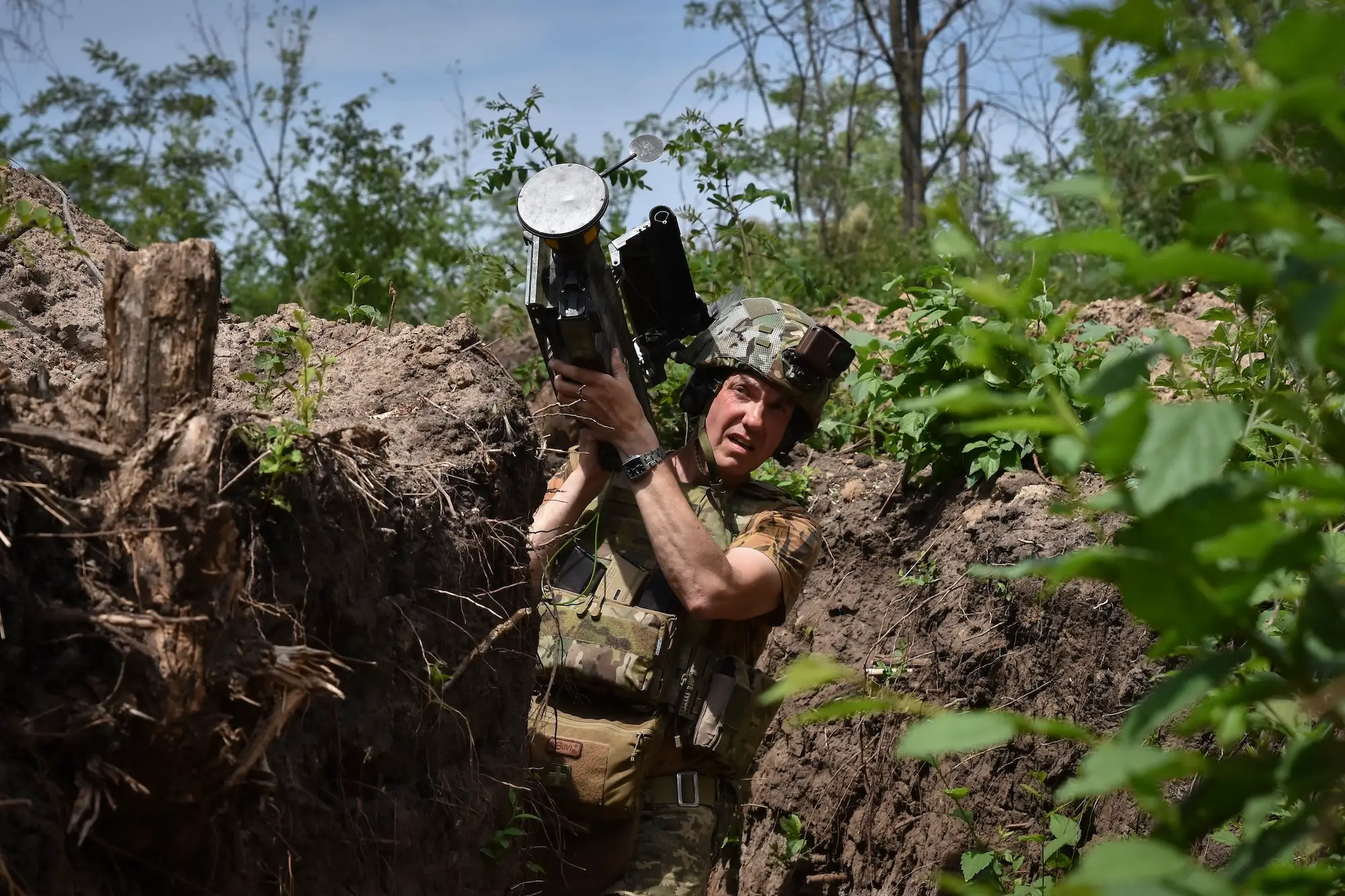 Camouflaged soldier holds a shoulder-fired launcher while standing in an earthen trench surrounded by dense greenery.