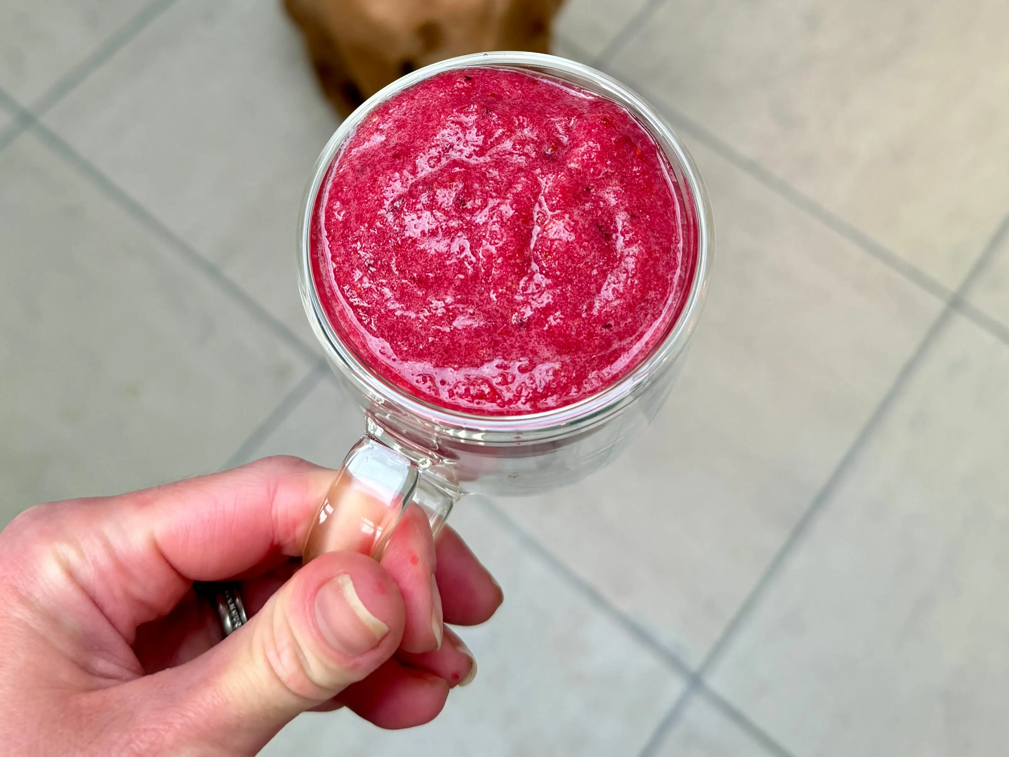 A brown dog sits on a tiled kitchen floor behind a hand holding a glass cup of bright red smoothie.