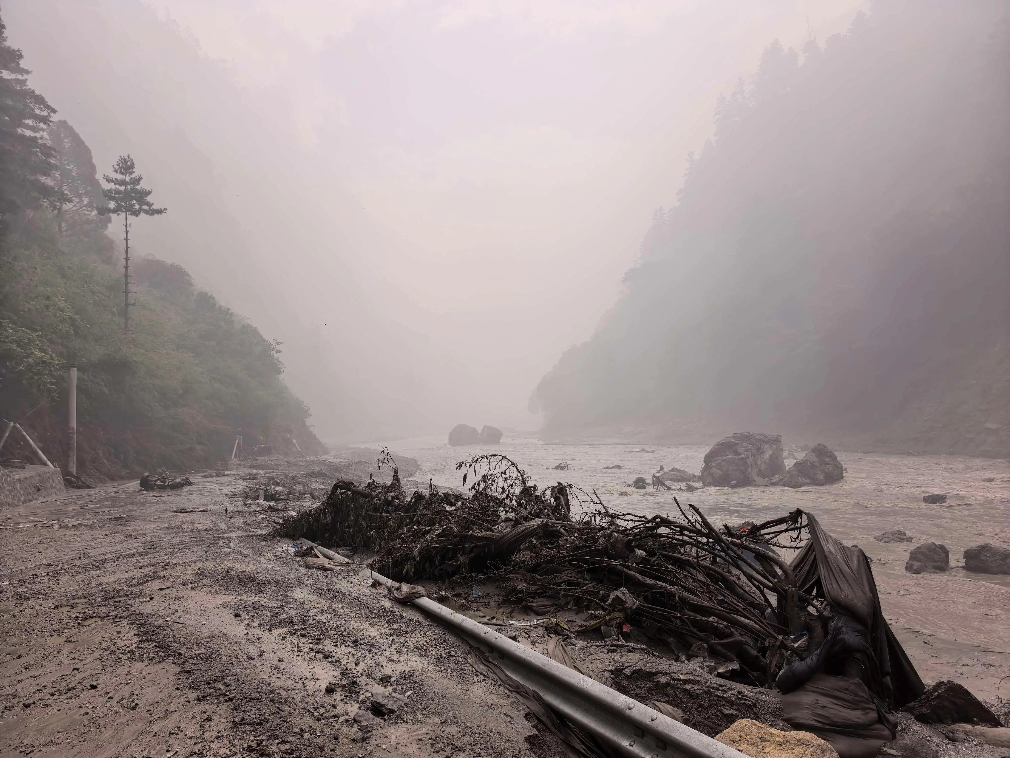 Mud, debris, and a damaged guardrail line a foggy riverside road in a steep forested canyon.