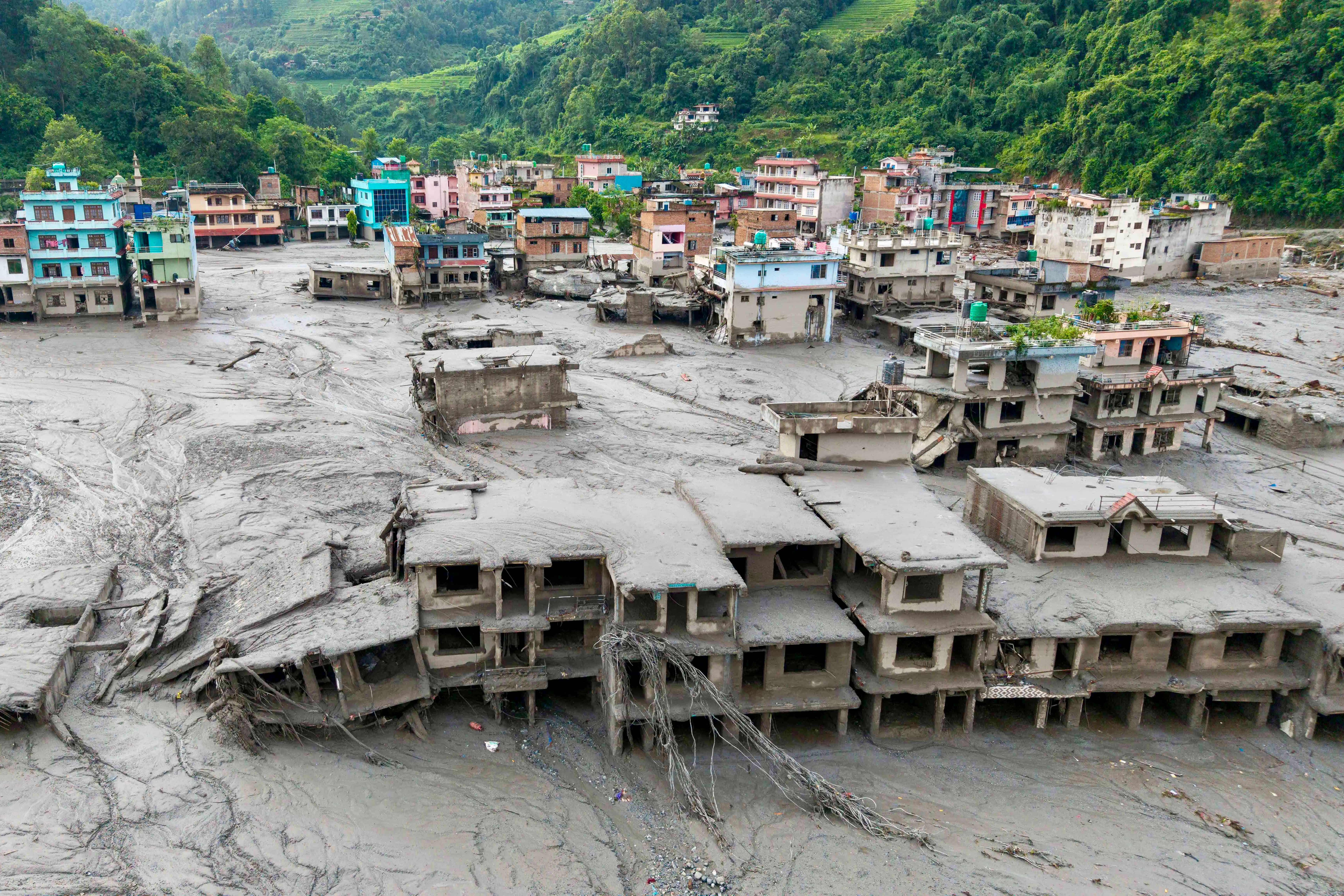 Buildings in a mountain valley town are surrounded and partly buried by thick gray mud and debris.