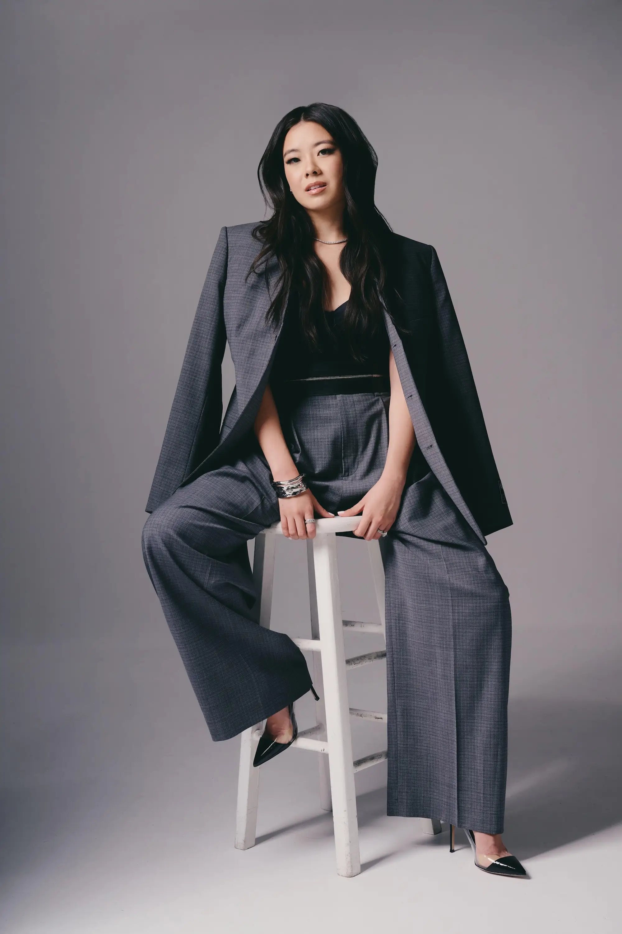 Person with long dark hair sits on a white stool wearing a gray suit against a gray studio backdrop.