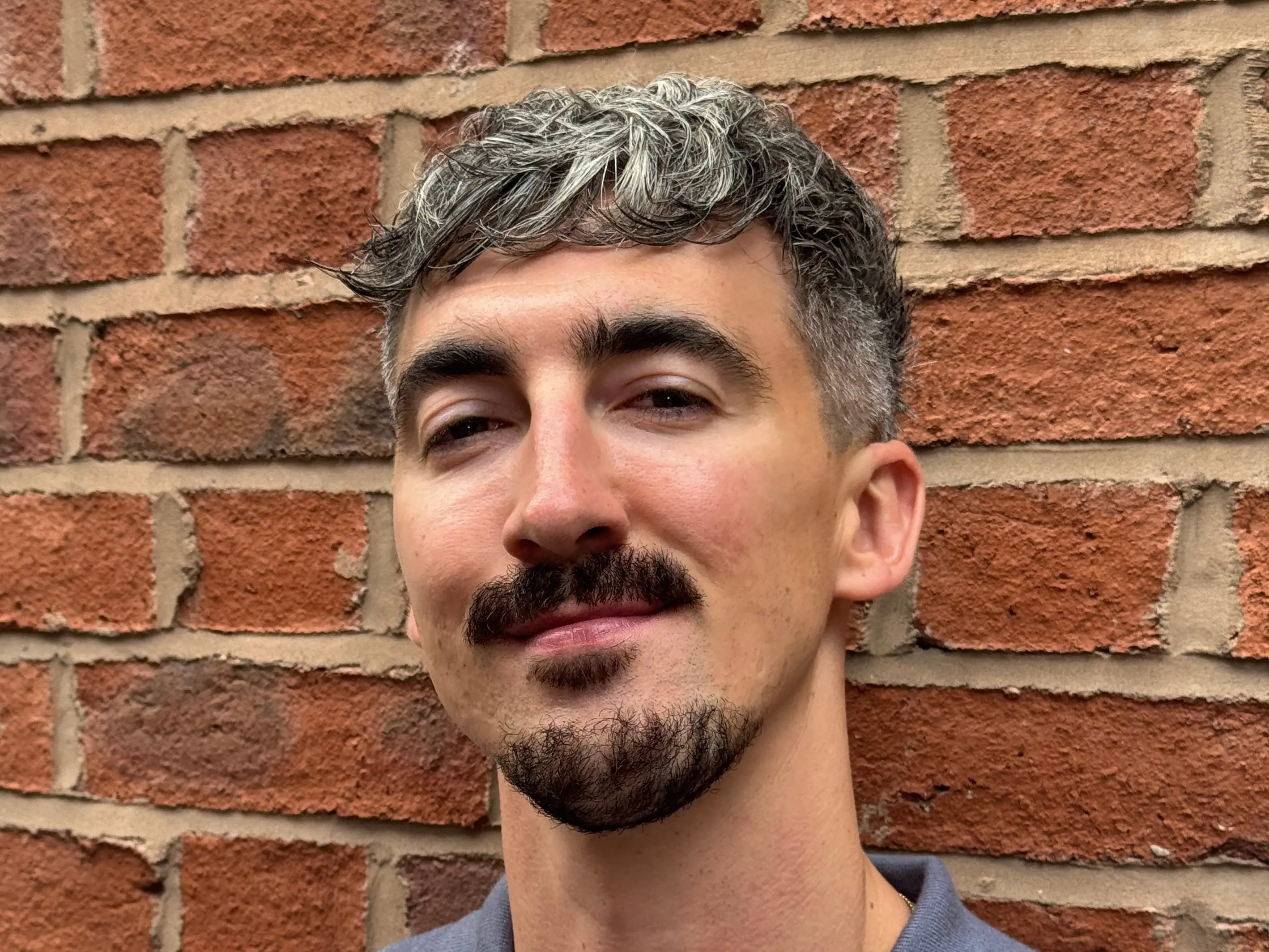 Person with short wavy hair and facial hair wearing a blue polo shirt against a brick wall.