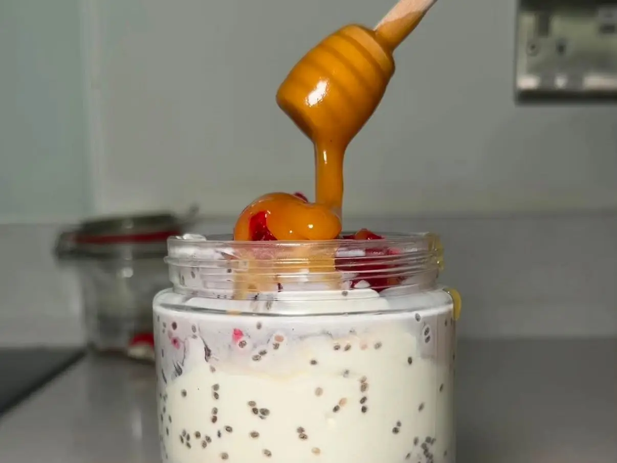 Honey drizzles from a wooden dipper onto a glass jar of creamy chia seed breakfast with red fruit.
