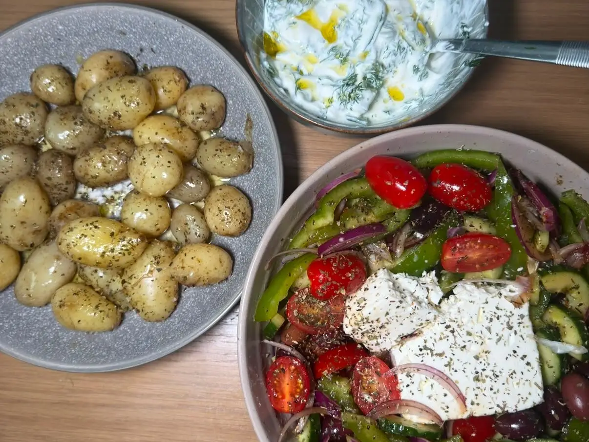 Overhead view of potatoes, Greek-style salad with feta, olives and vegetables, and a bowl of yogurt herb sauce.