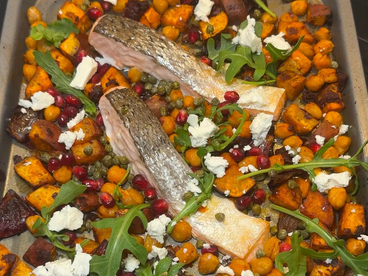 Roasted fish fillets sit on a tray with chickpeas, orange vegetable cubes, greens, pomegranate seeds, capers, and white cheese.