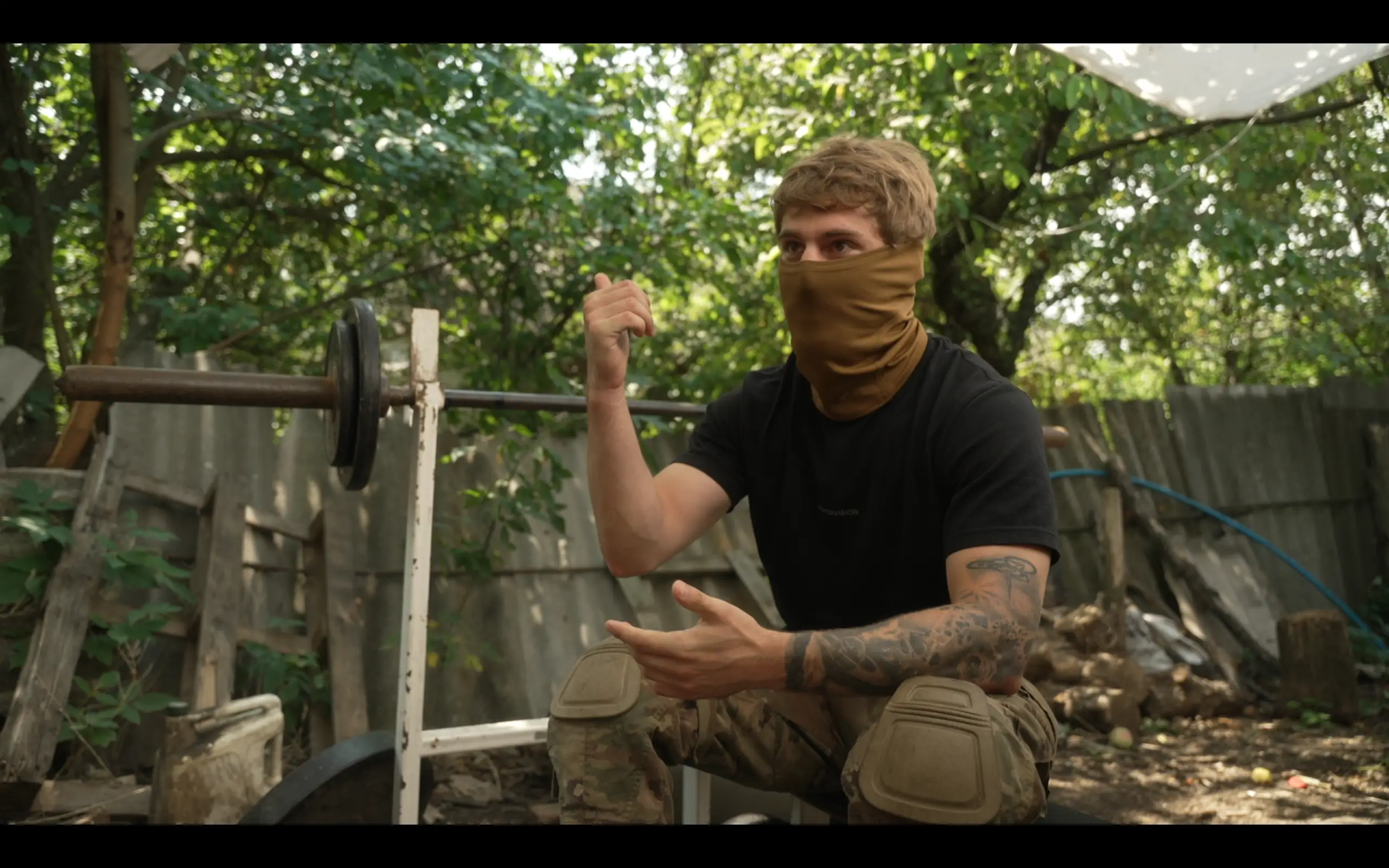 Masked person in camouflage pants gestures while seated beside an outdoor barbell bench in a wooded yard.