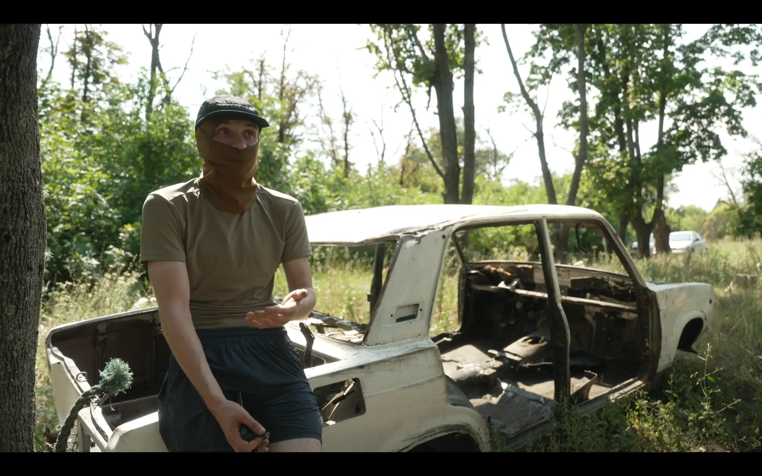 Masked person sits on a stripped, burned-out car in a wooded roadside area with recording equipment nearby.