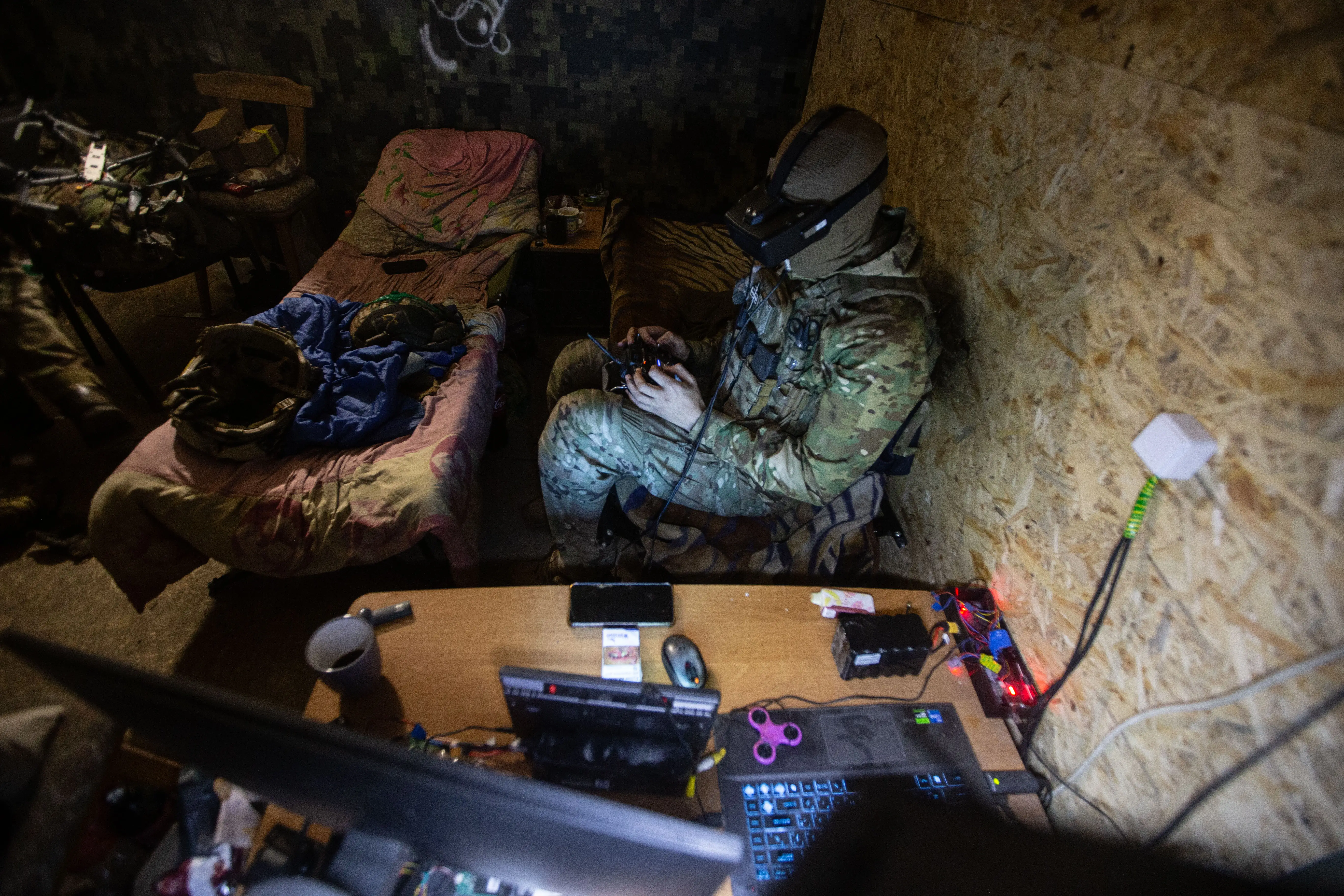 Camouflage-clad person with tactical gear sits at a desk with laptop and electronics in a dim room.
