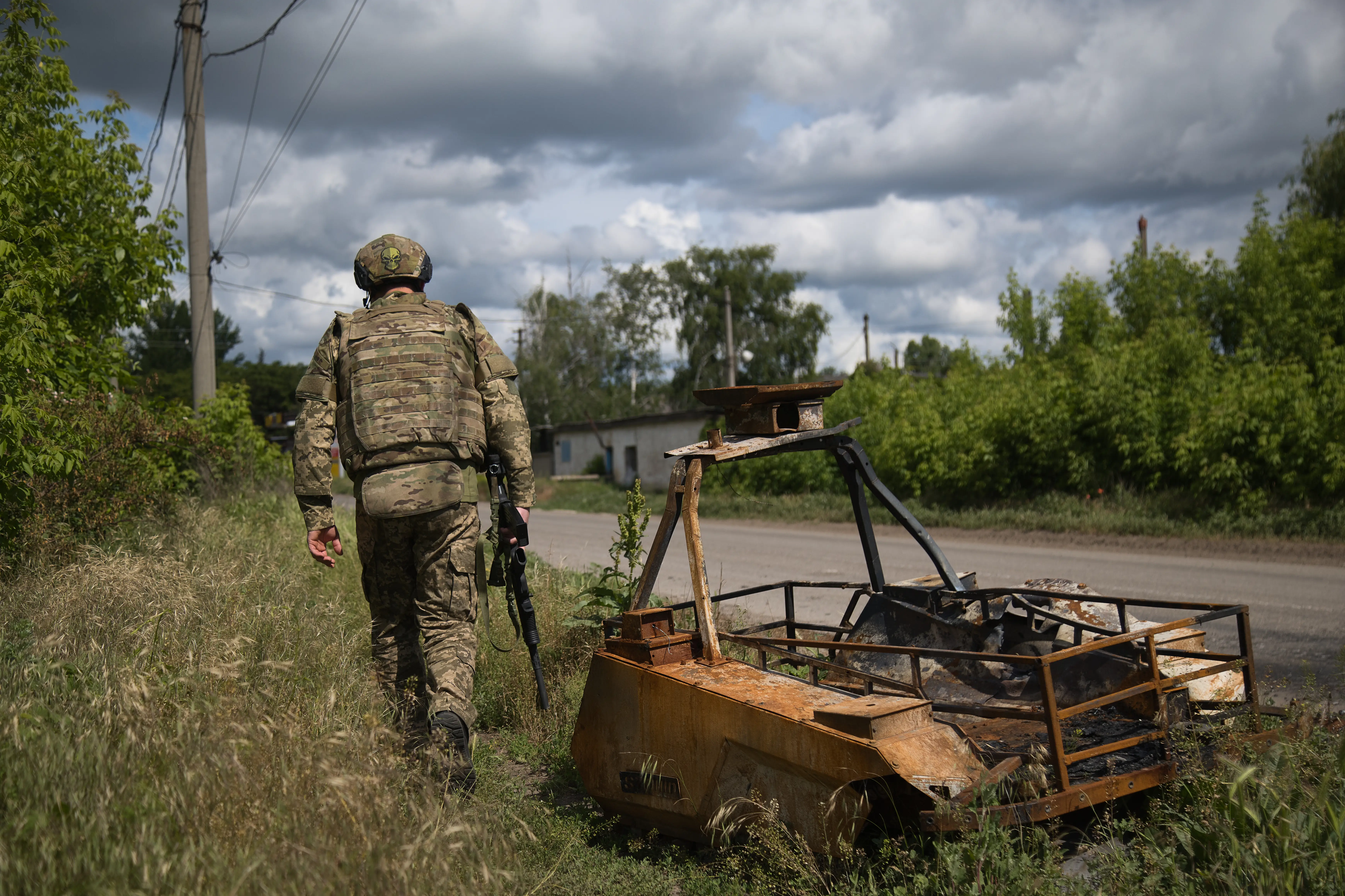 Camouflaged soldier walks along a rural roadside beside a burned-out vehicle under a cloudy sky.