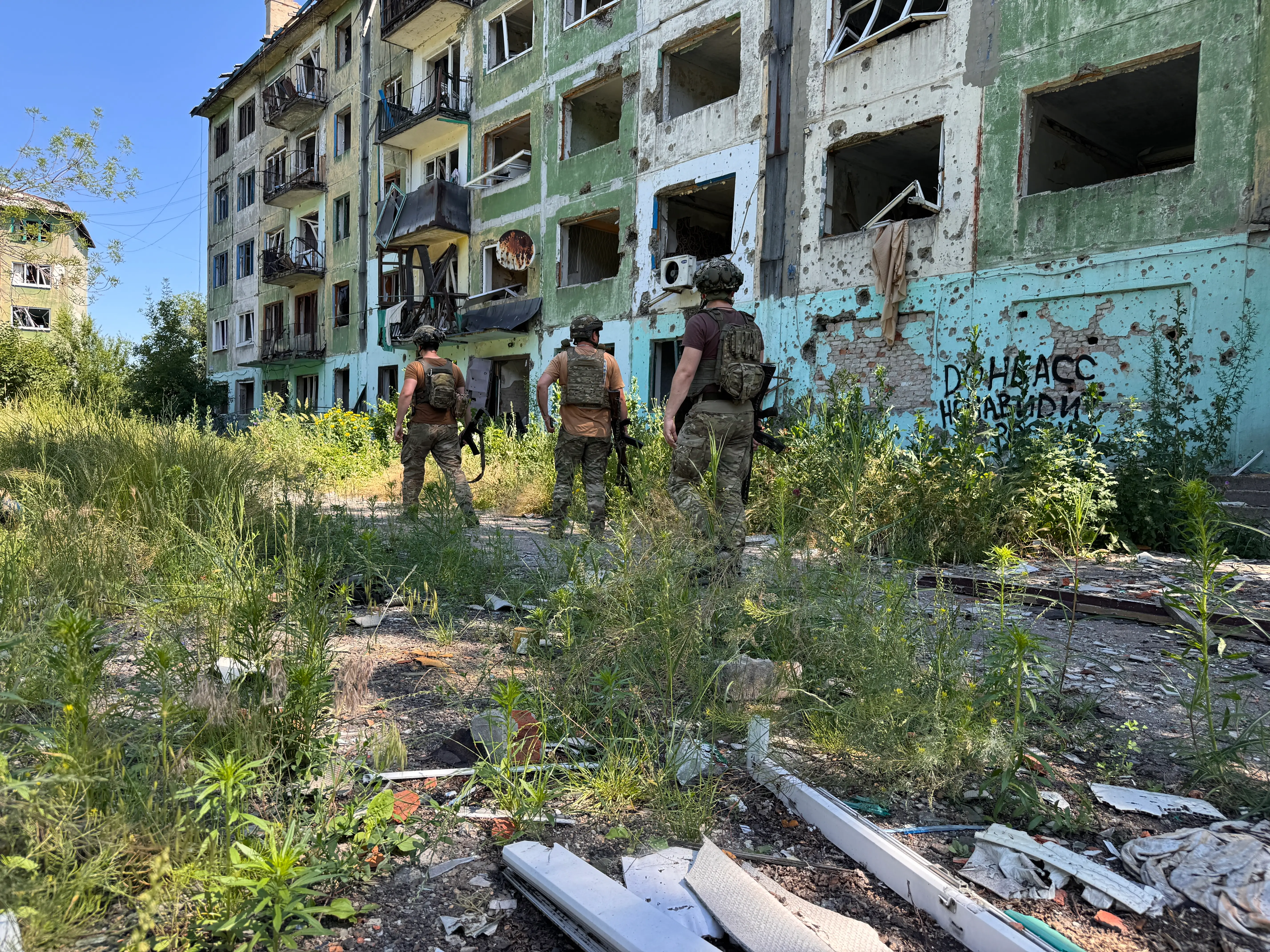 Three uniformed personnel walk through overgrown debris beside a heavily damaged apartment building with broken windows.