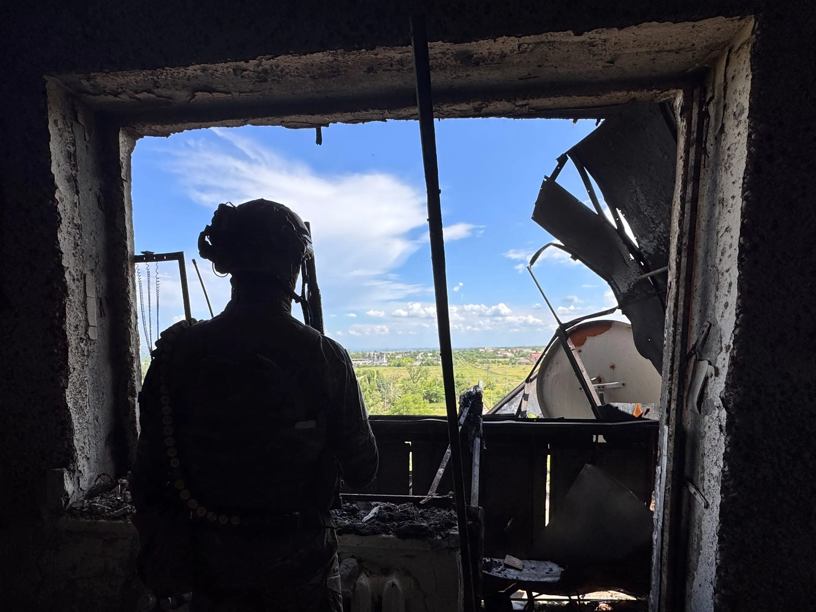 Silhouetted person stands inside a damaged room looking out through a broken window toward a bright landscape.