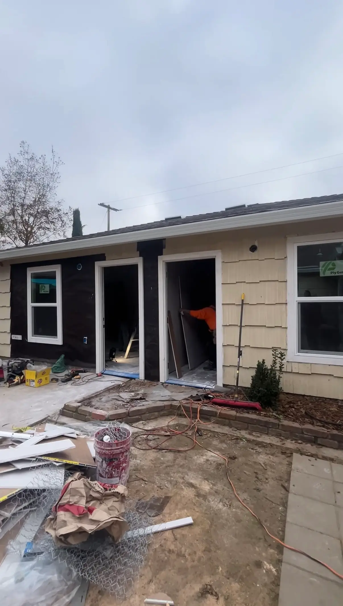 Single-story house exterior undergoing renovation with open doorways, tools, debris, and a partially visible worker inside.