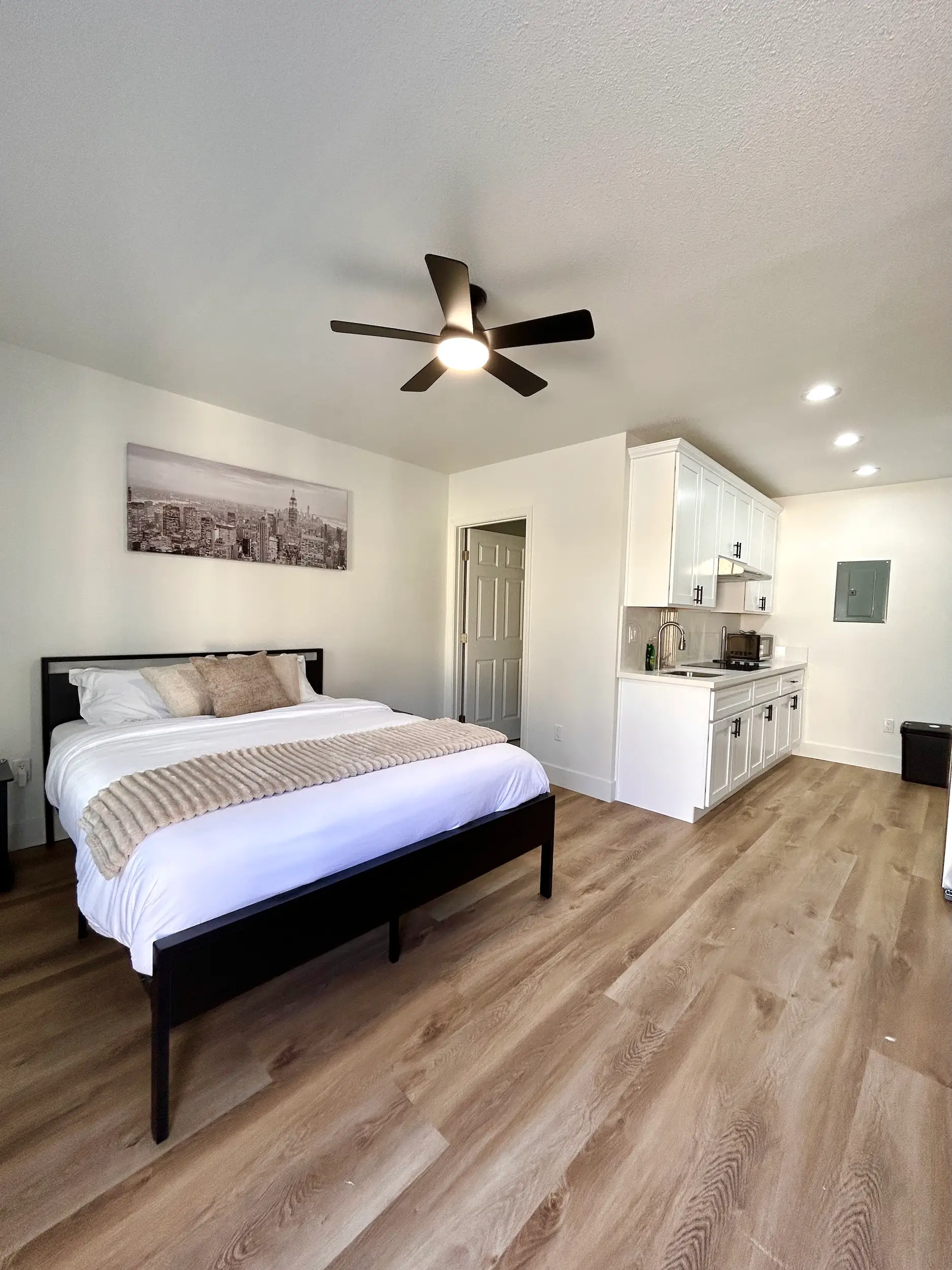 Modern studio room with a made bed, ceiling fan, kitchenette, wood-look flooring, and neutral decor.