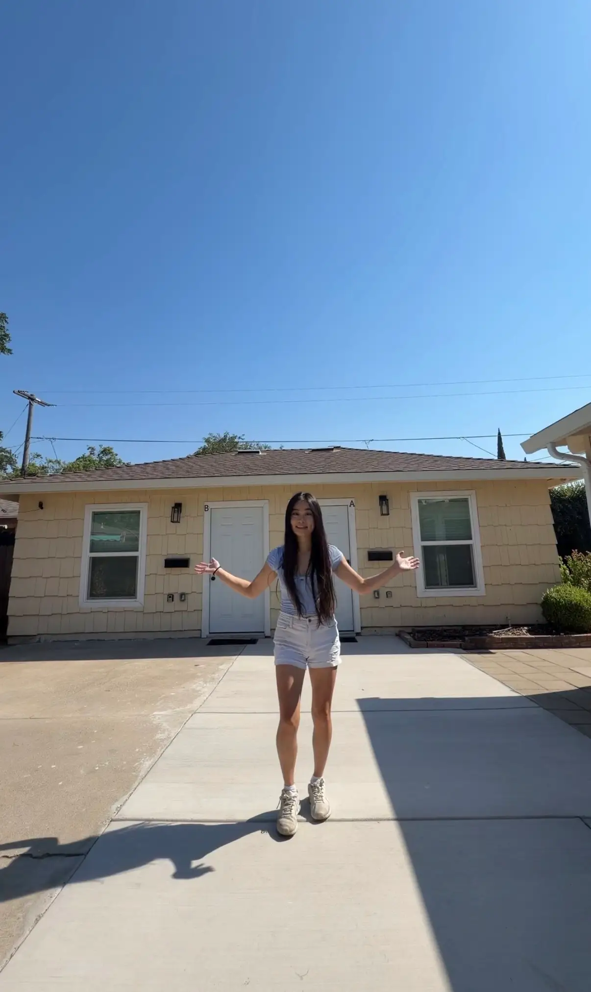 Person stands smiling with arms out on a driveway in front of a beige two-door residential building.