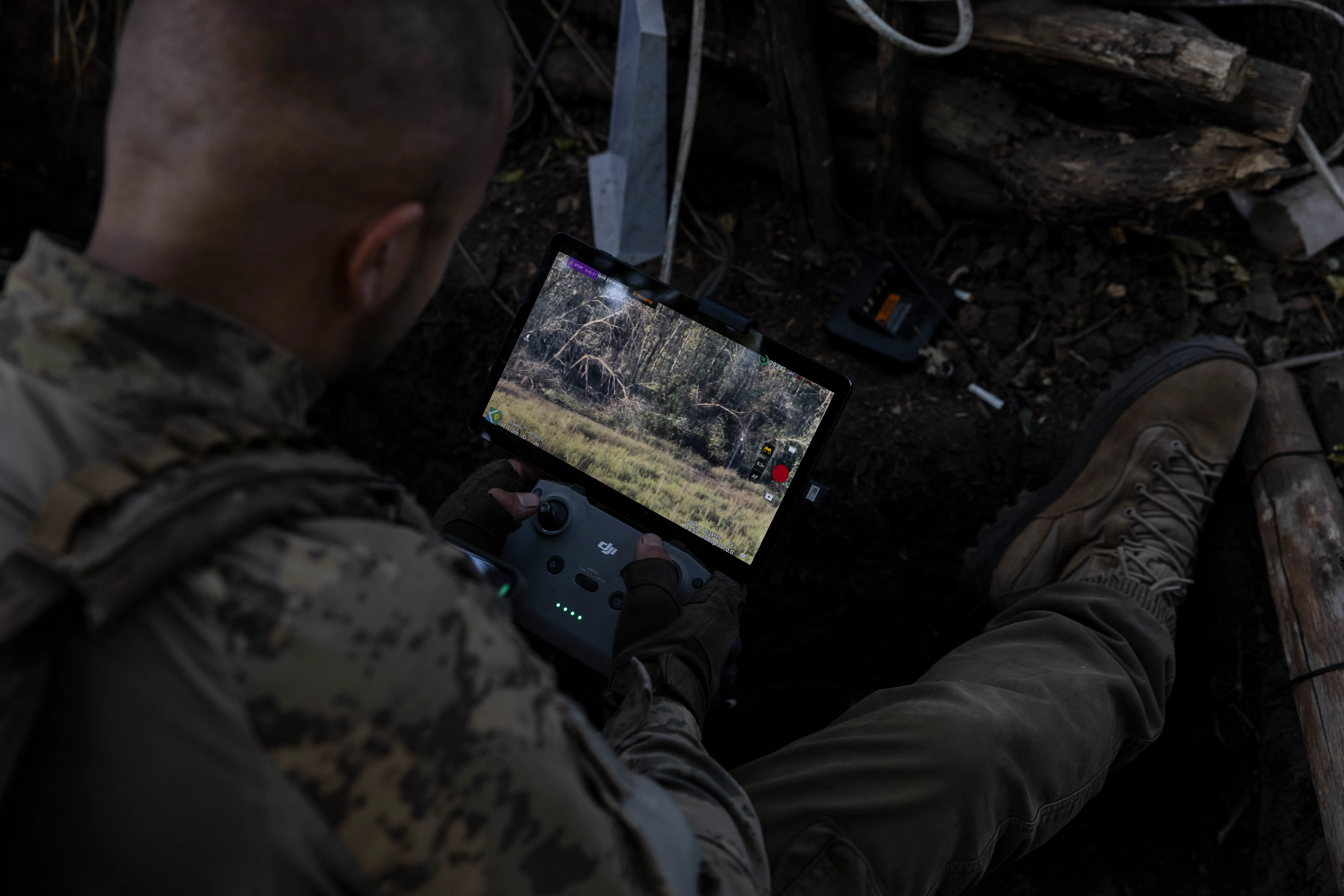 A person in camouflage clothing operates a screen-equipped controller in a dark outdoor dugout or wooded position.